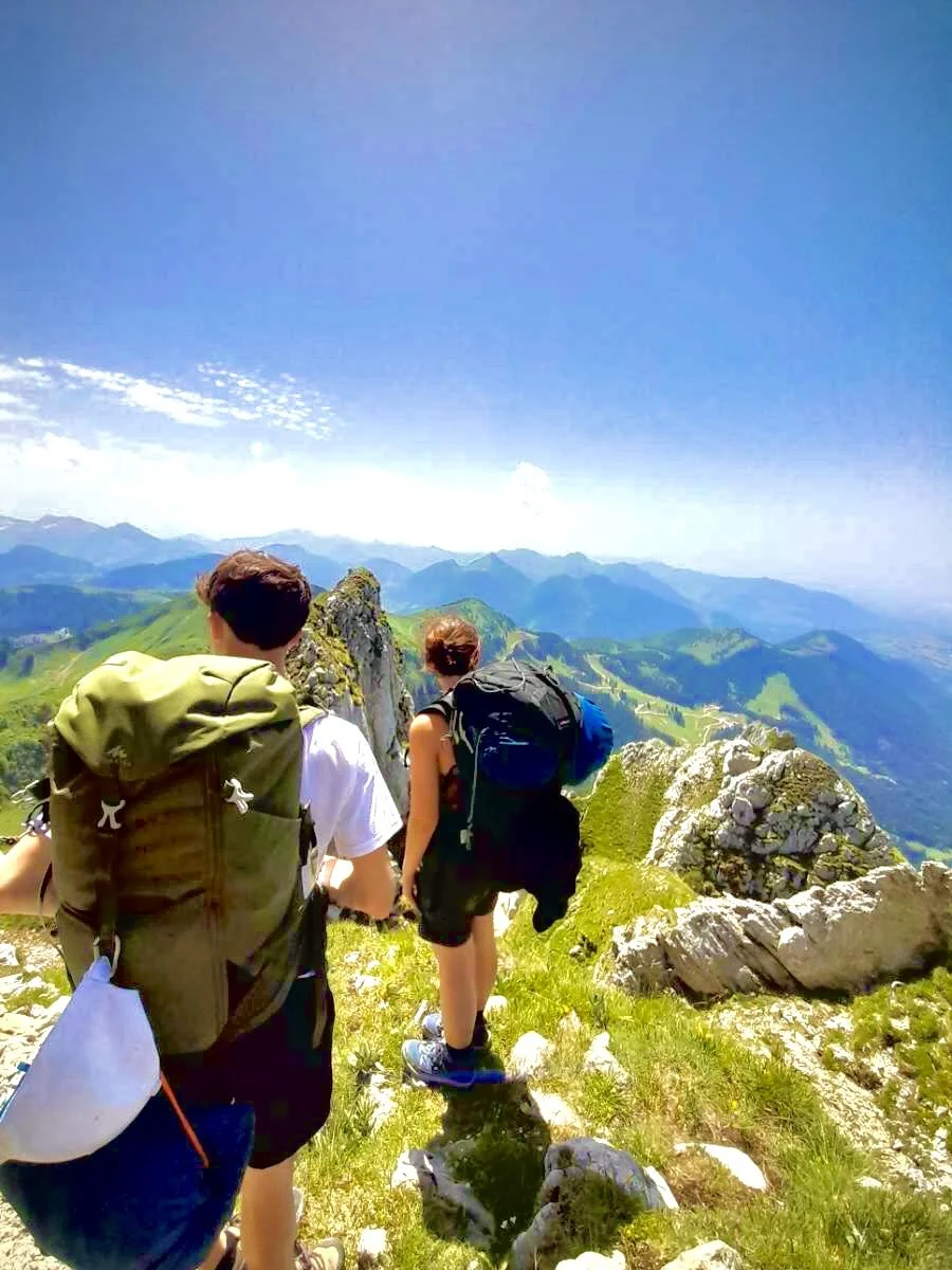 Deux randonneurs avec des sacs à dos marchant sur un sentier en montagne, avec des sommets et un ciel bleu en arrière-plan.