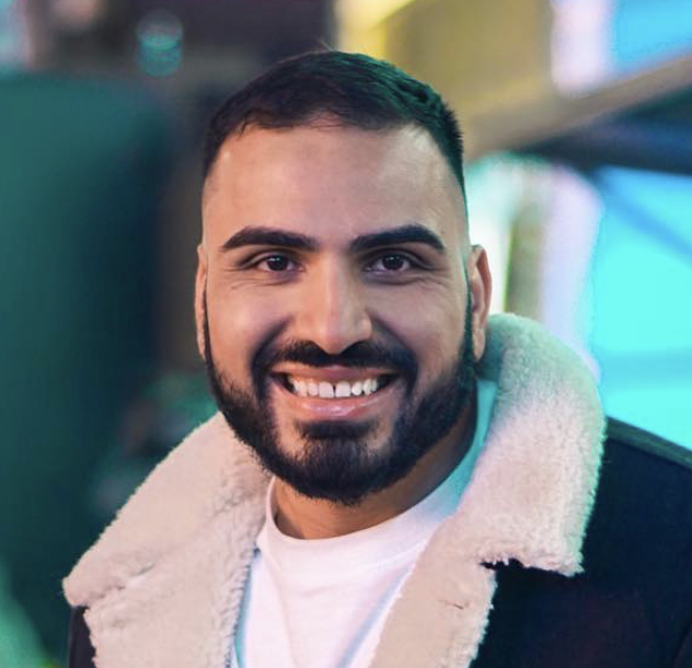 A man with dark, curly hair, glasses, a nose ring, and a beard, wearing a dark sweater, sitting indoors with a plant in the background.