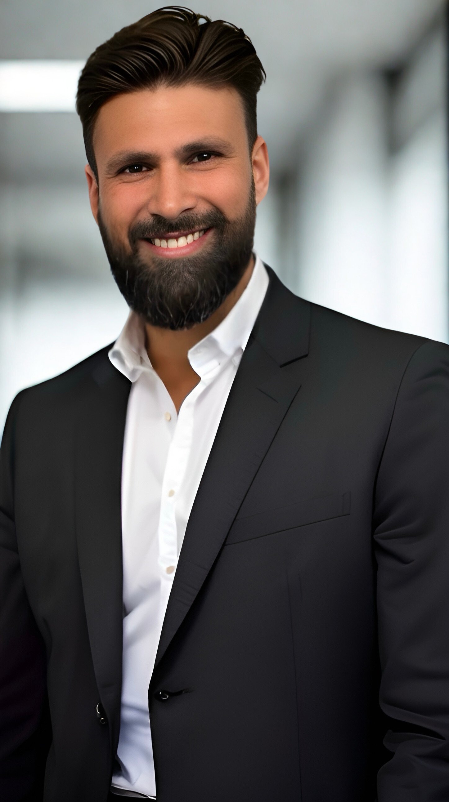 A smiling man with dark hair, a full beard, wearing a black suit and white shirt, standing in a professional office environment.