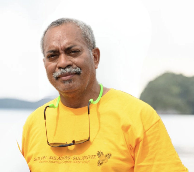 A middle-aged man with gray hair and a mustache, wearing a bright yellow shirt and sunglasses hanging around his neck, standing outdoors near a body of water with a blurred landmass in the background.