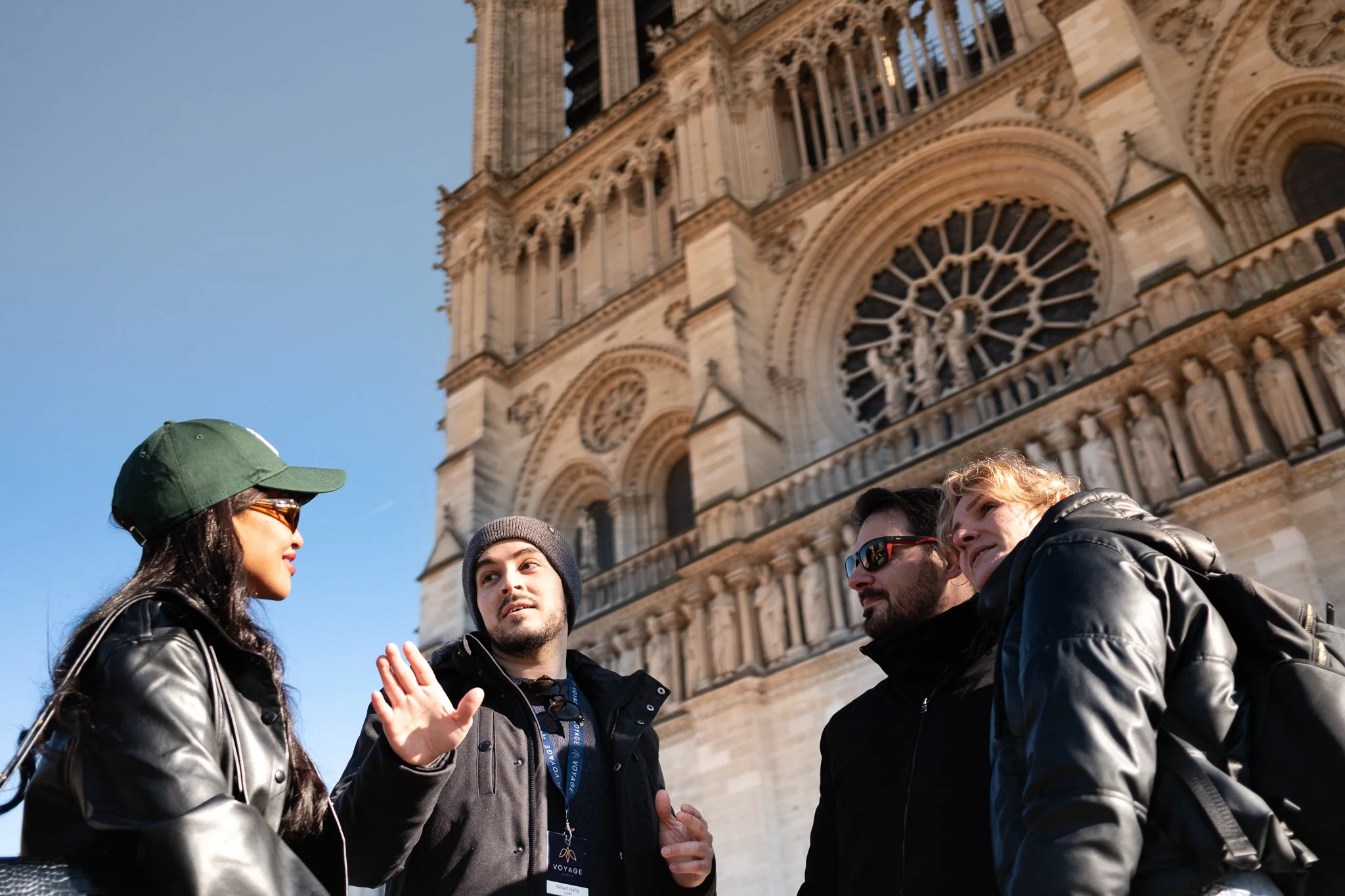 Four people, wearing winter clothes, stand in front of the Notre-Dame Cathedral in Paris, engaging in conversation. The gothic architecture and intricate details of the cathedral are visible in the background.