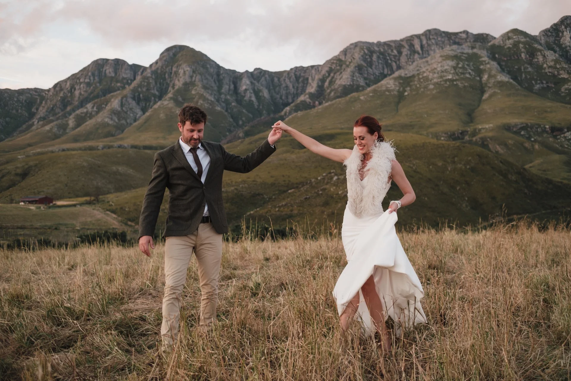 A couple dancing in a grassy field with mountains in the background during sunset.