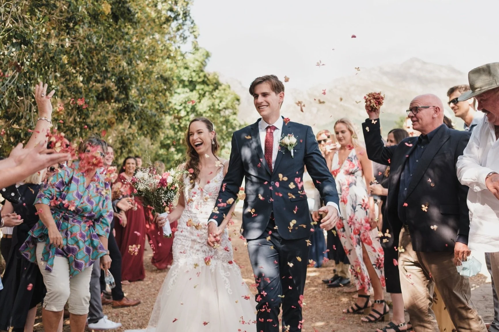 Happy bride and groom holding hands, walking through a celebration crowd with confetti and flower petals, outdoors on a sunny day.