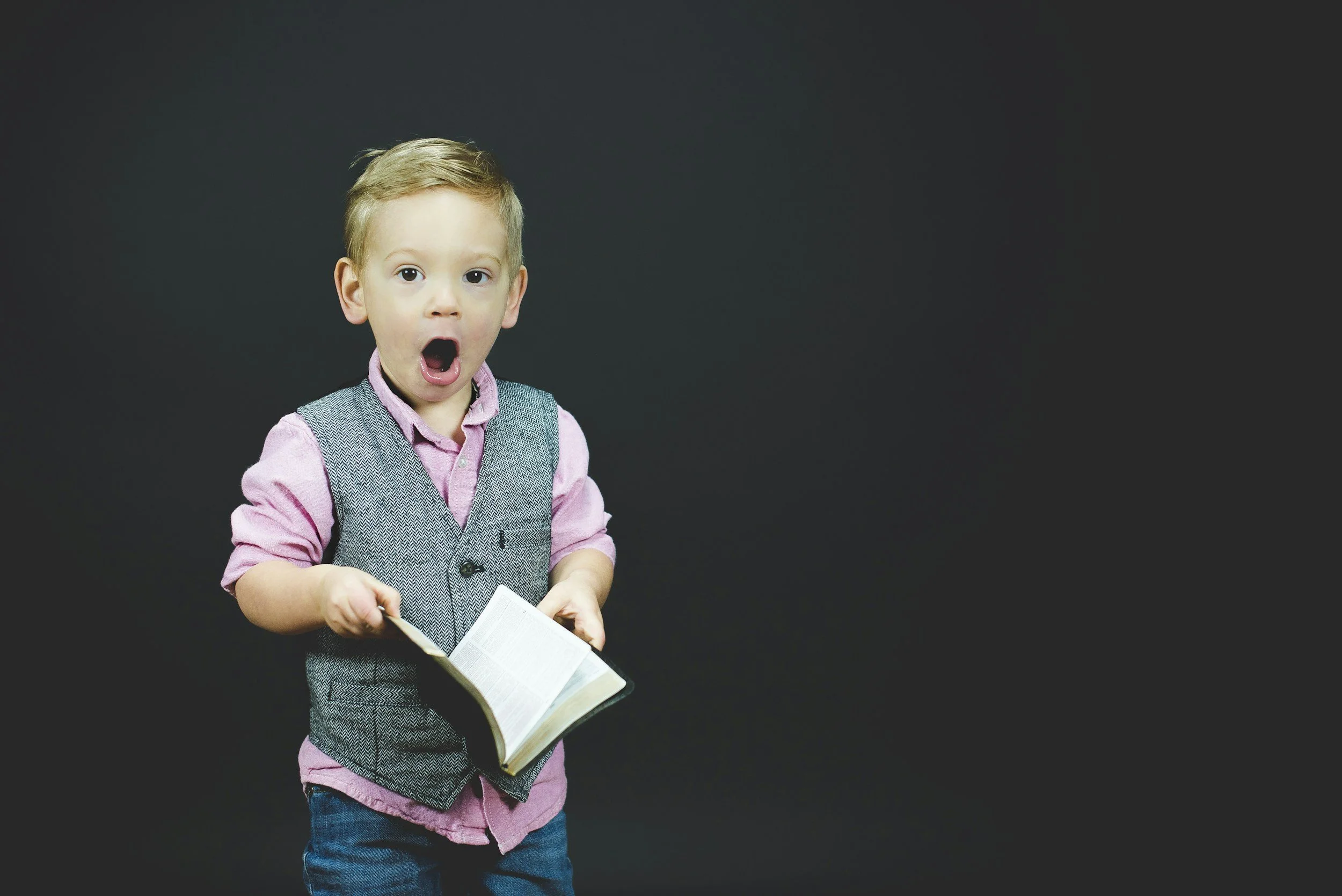 A young boy with blond hair, wearing a pink shirt and gray vest, holding an open book with a surprised expression, against a black background.