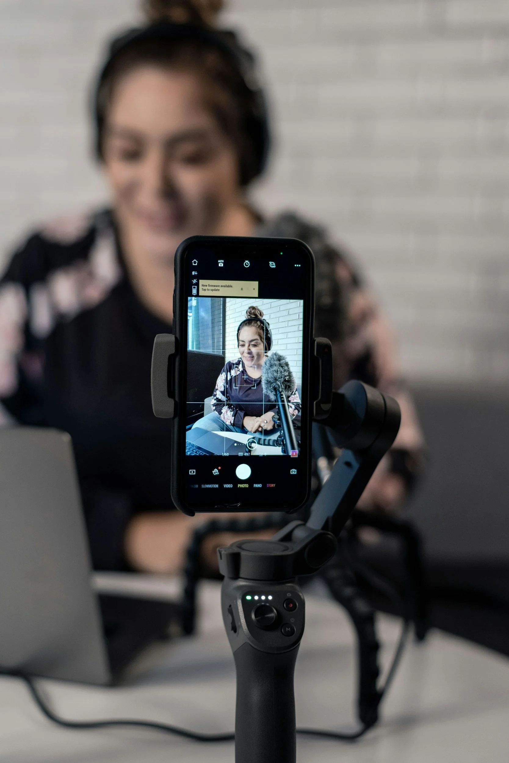 A person is being recorded with a smartphone mounted on a tripod. The phone screen shows a woman with dark hair in a bun, wearing a floral top and smiling. The background has a light-colored brick wall.