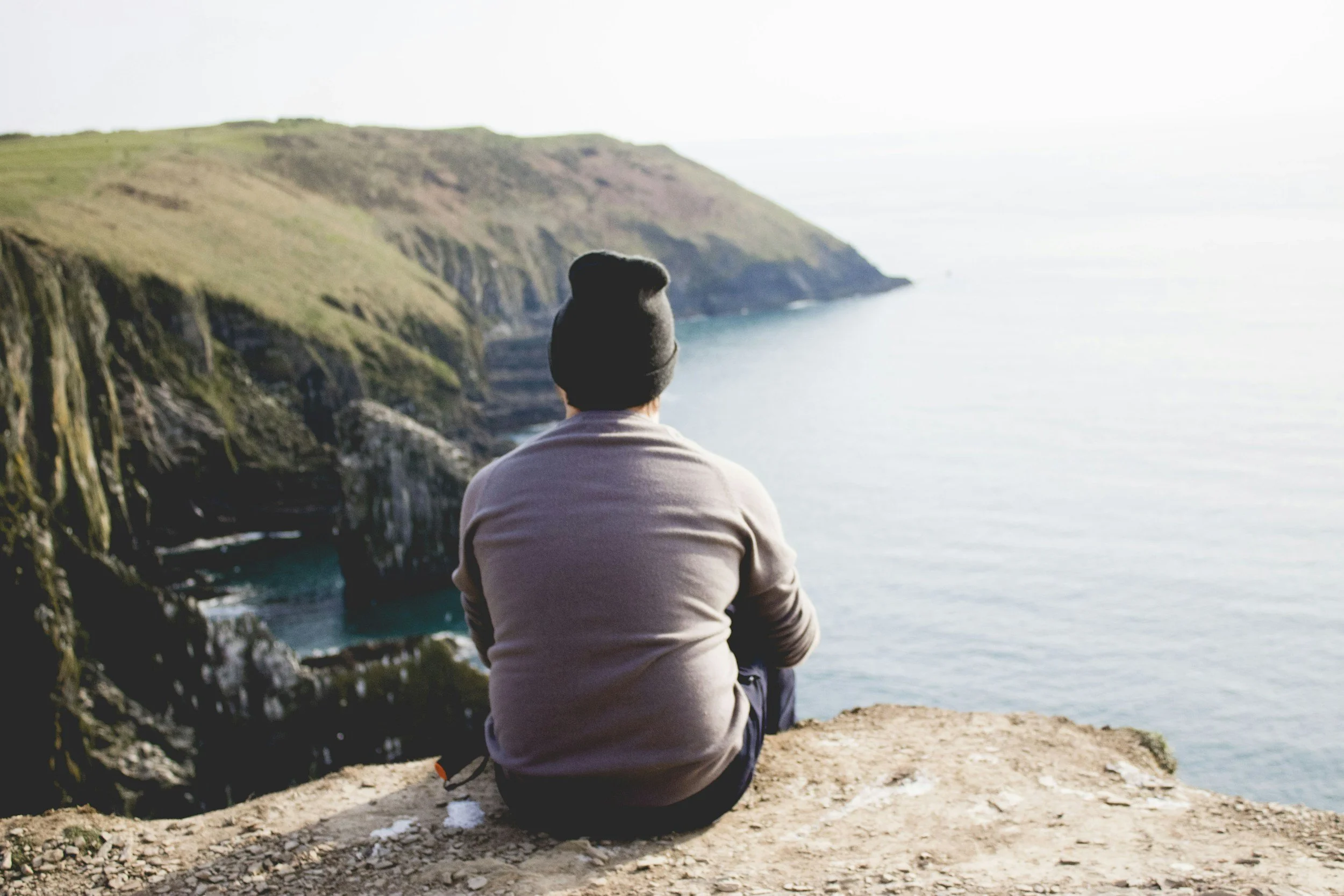 A person wearing a beanie hat and a beige jacket sitting on a rocky cliff, looking out over the ocean with green cliffs in the distance.