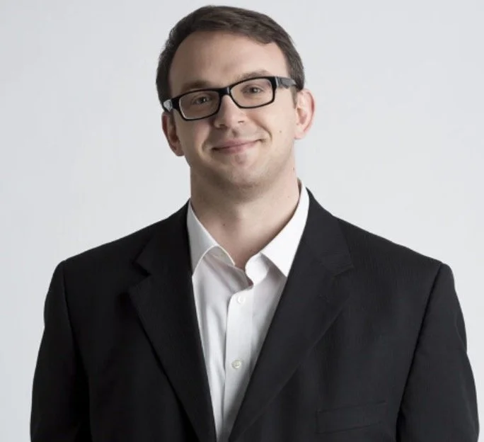 A smiling man in a suit with no tie, wearing glasses