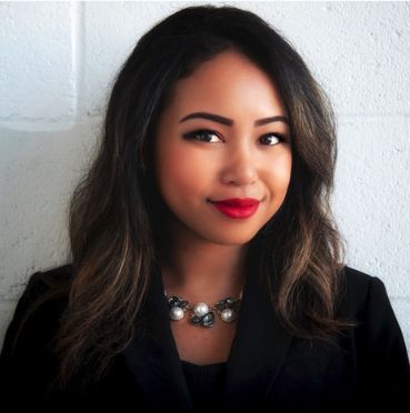 A woman with long dark hair, red lipstick, wearing a black blazer and a statement necklace, standing against a white wall.