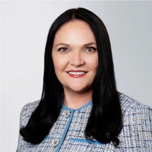 Portrait of a woman with long dark hair wearing a light gray blazer with a zipper detail, smiling at the camera.