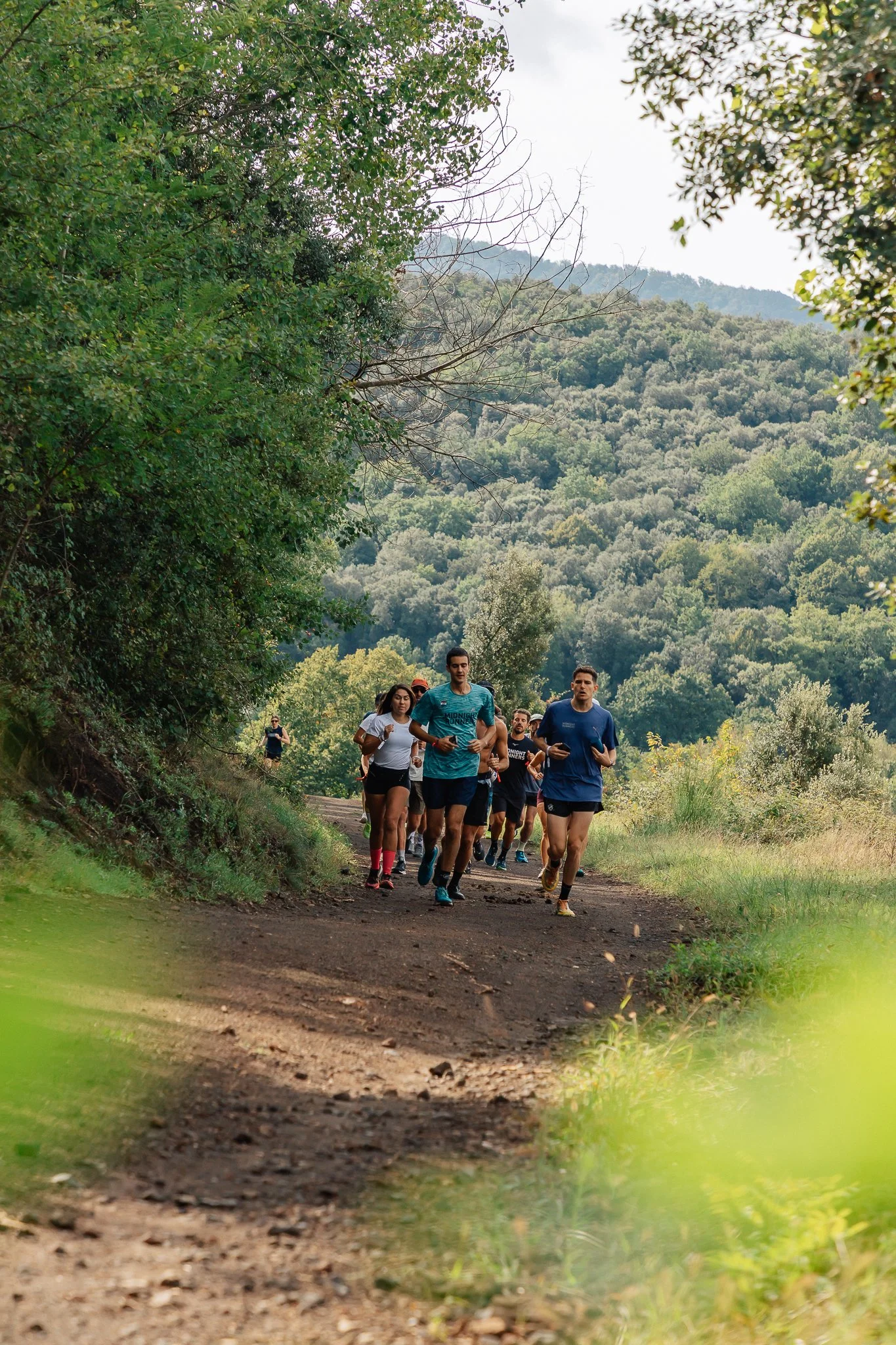 Group of people jogging on a trail surrounded by trees and grass, under a blue sky.