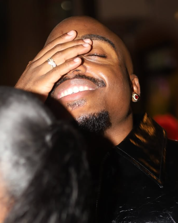 A man smiling and laughing with his eyes closed, holding his hand over his face, showing an engagement ring. He has earrings and facial hair, and is wearing a black jacket with a shiny collar.