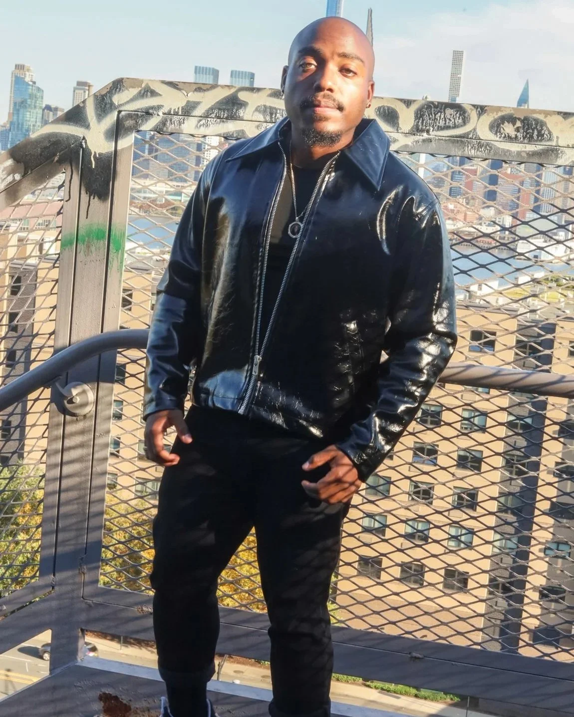 A man wearing a shiny black leather jacket and black pants stands on a rooftop with a metal guardrail, with a city skyline in the background during daytime.