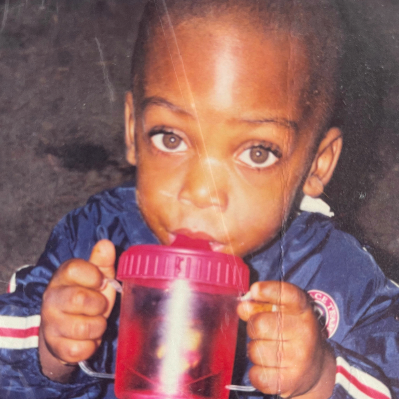 A young child with curly dark hair, brown eyes, and wearing a blue jacket is holding a red container and looking at the camera.