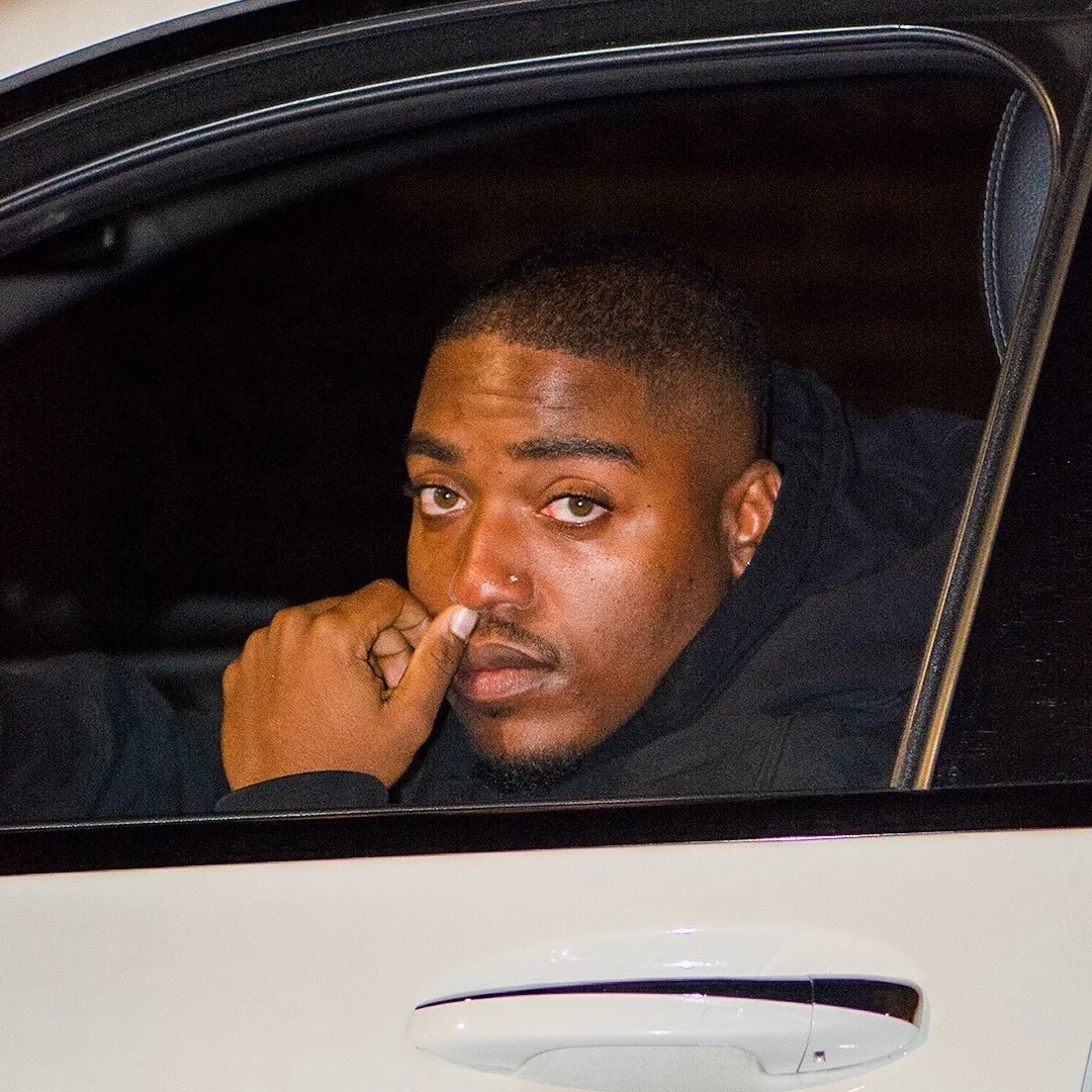 A young man with short hair and a nose piercing sitting in a car, looking out the window and touching his nose.