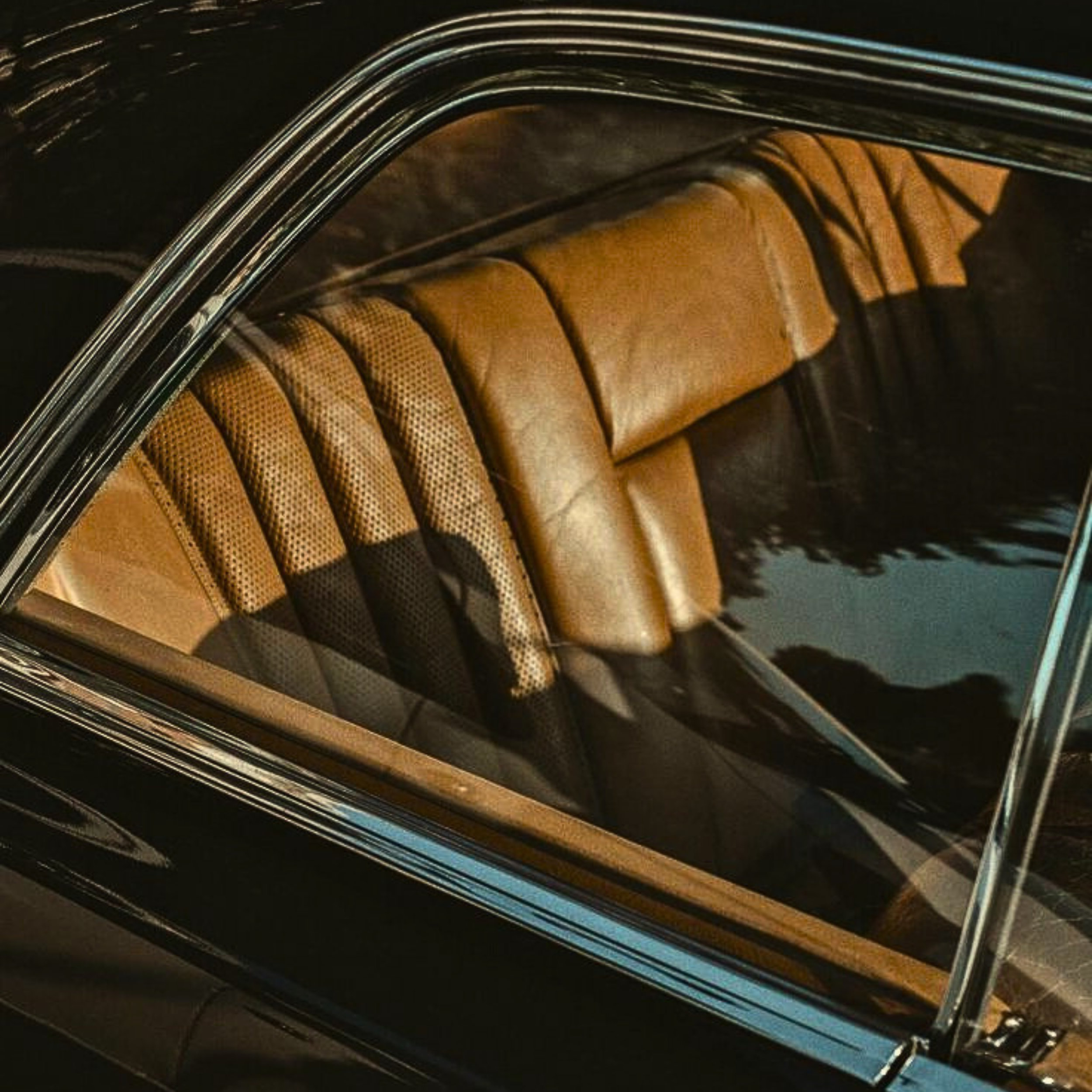 Close-up of the tan leather interior of a vintage car seen through the window, with reflections of trees on the glass.