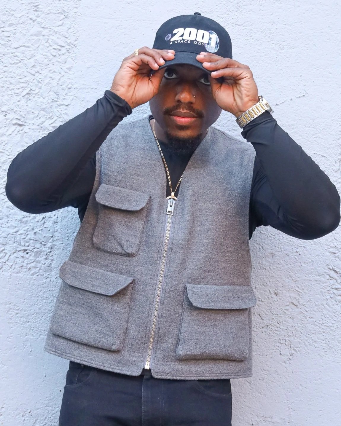 A man wearing a black cap, gray vest, black long-sleeve shirt, and a wristwatch, posing against a textured white wall.