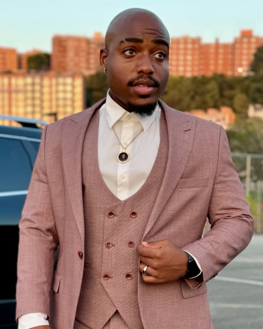 Man in a pink suit with a white shirt and gold necklace standing outdoors near a parking lot with buildings in the background.