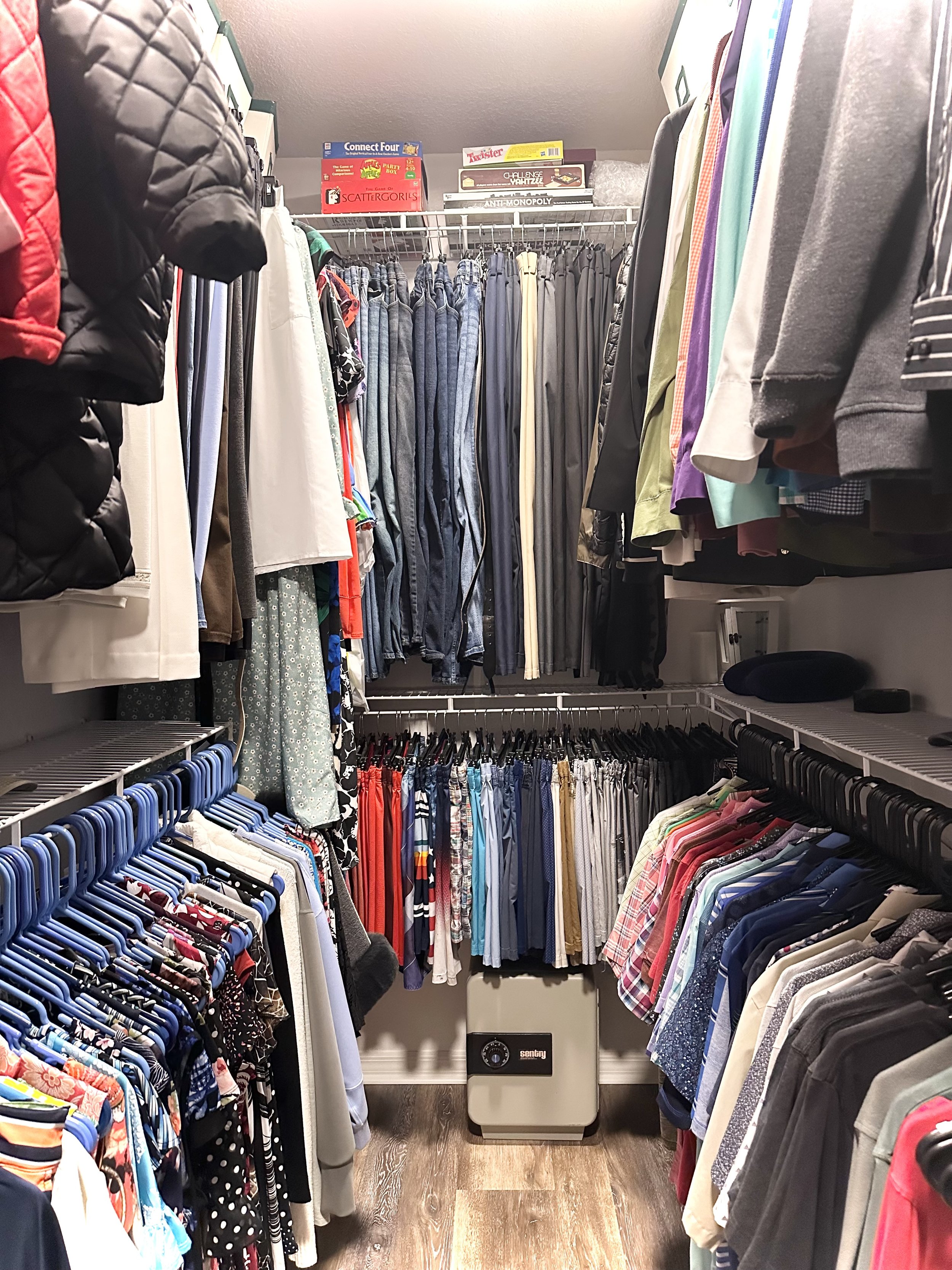 Organized closet with clothes on hangers, including shirts, pants, and jackets. Shelves hold board games like Connect Four and Scattergories. A security safe is on the floor.