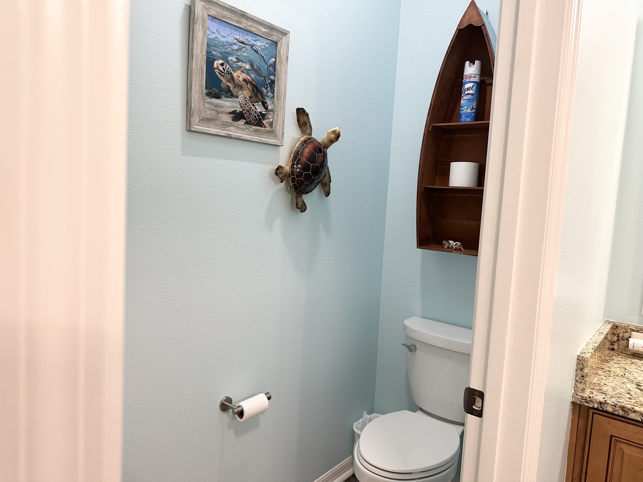 Bathroom with light blue walls, toilet paper holder, and wall decorations. A sea turtle picture and decorative turtle sculpture hang on the wall. A wooden shelf shaped like a boat holds Lysol spray and toilet paper. The counter features granite and w