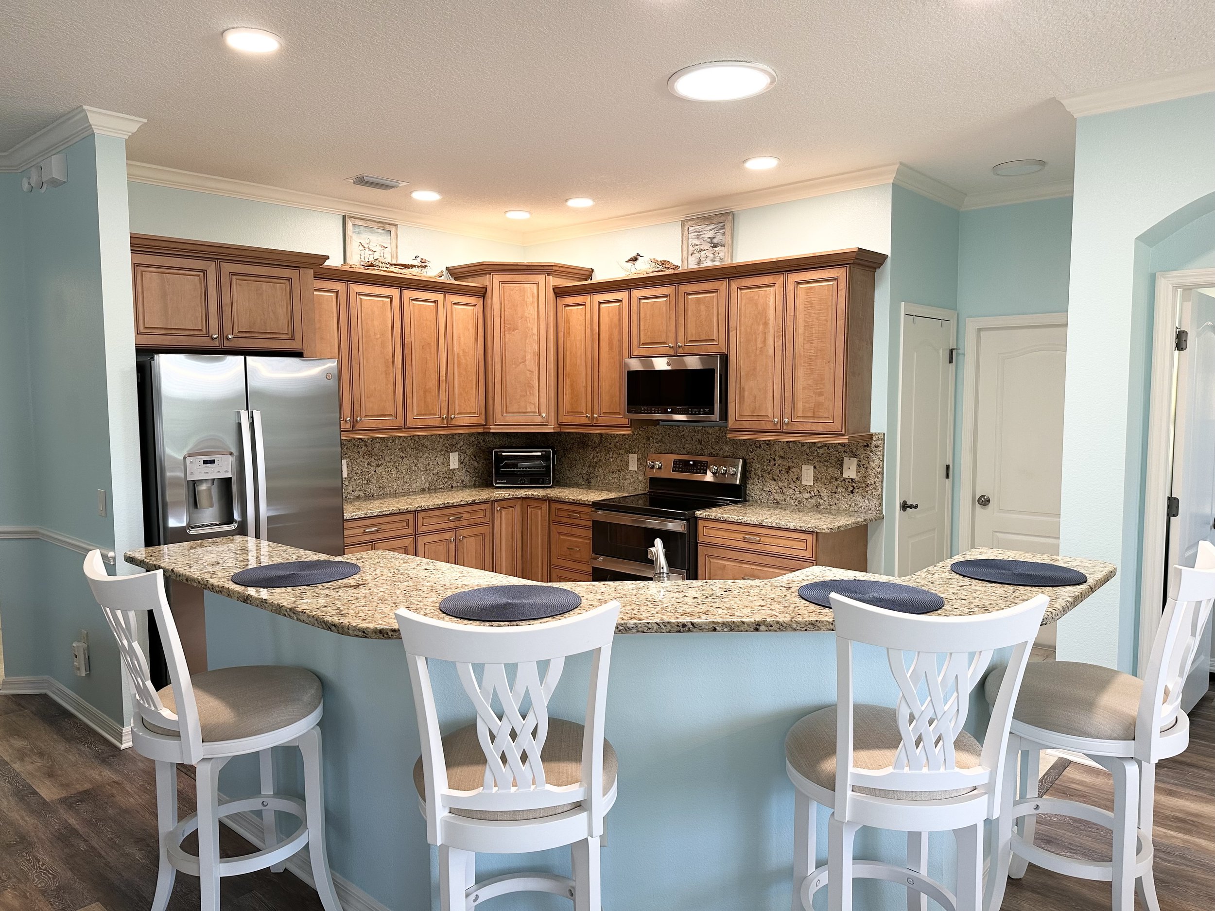Modern kitchen with light blue walls, granite countertops, wooden cabinets, stainless steel appliances, and white bar stools.