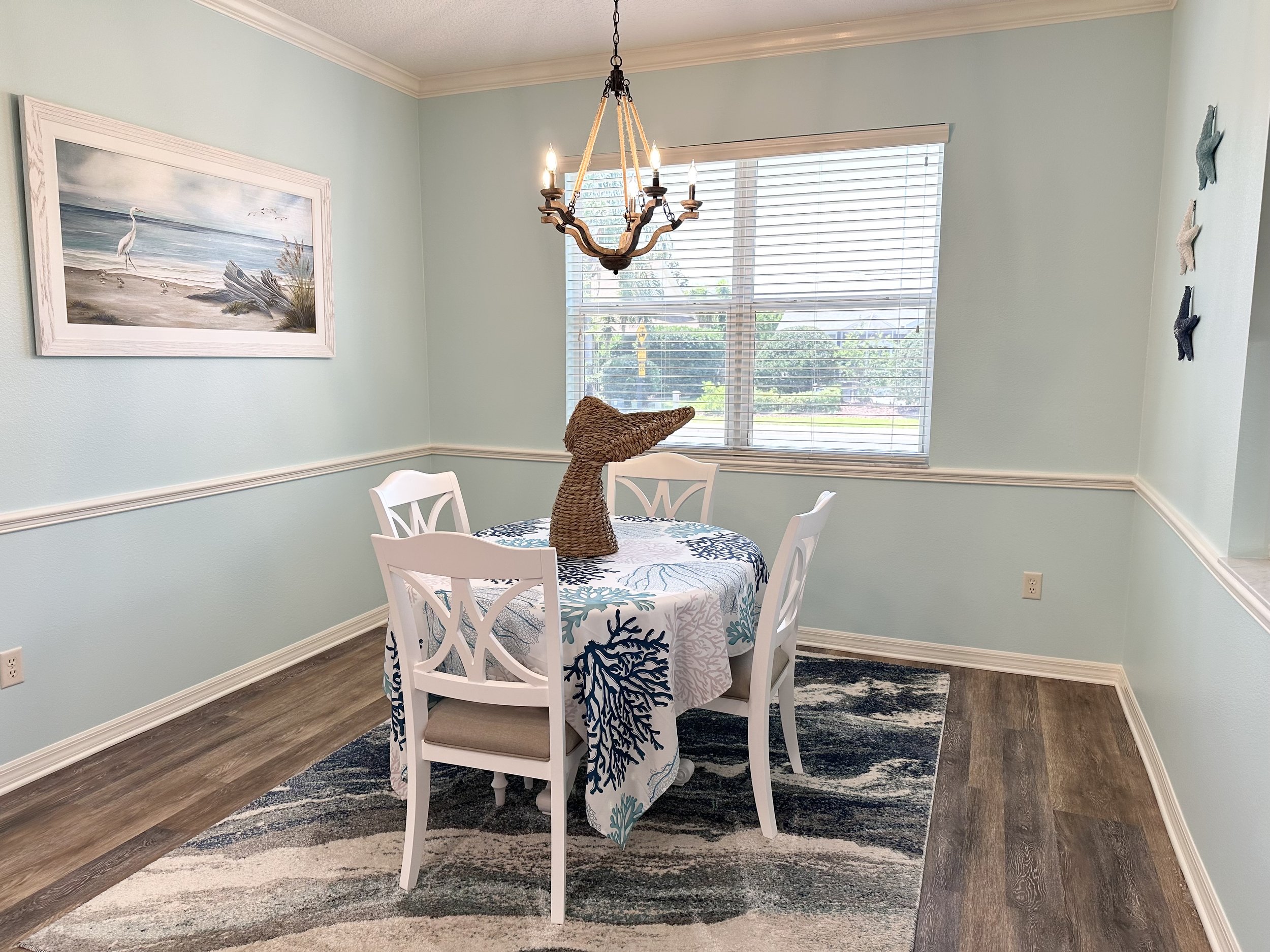 Coastal-themed dining room with light blue walls, a round table with a beach-patterned tablecloth, wicker dolphin centerpiece, white chairs, wall art of a seaside scene, and starfish wall decorations.