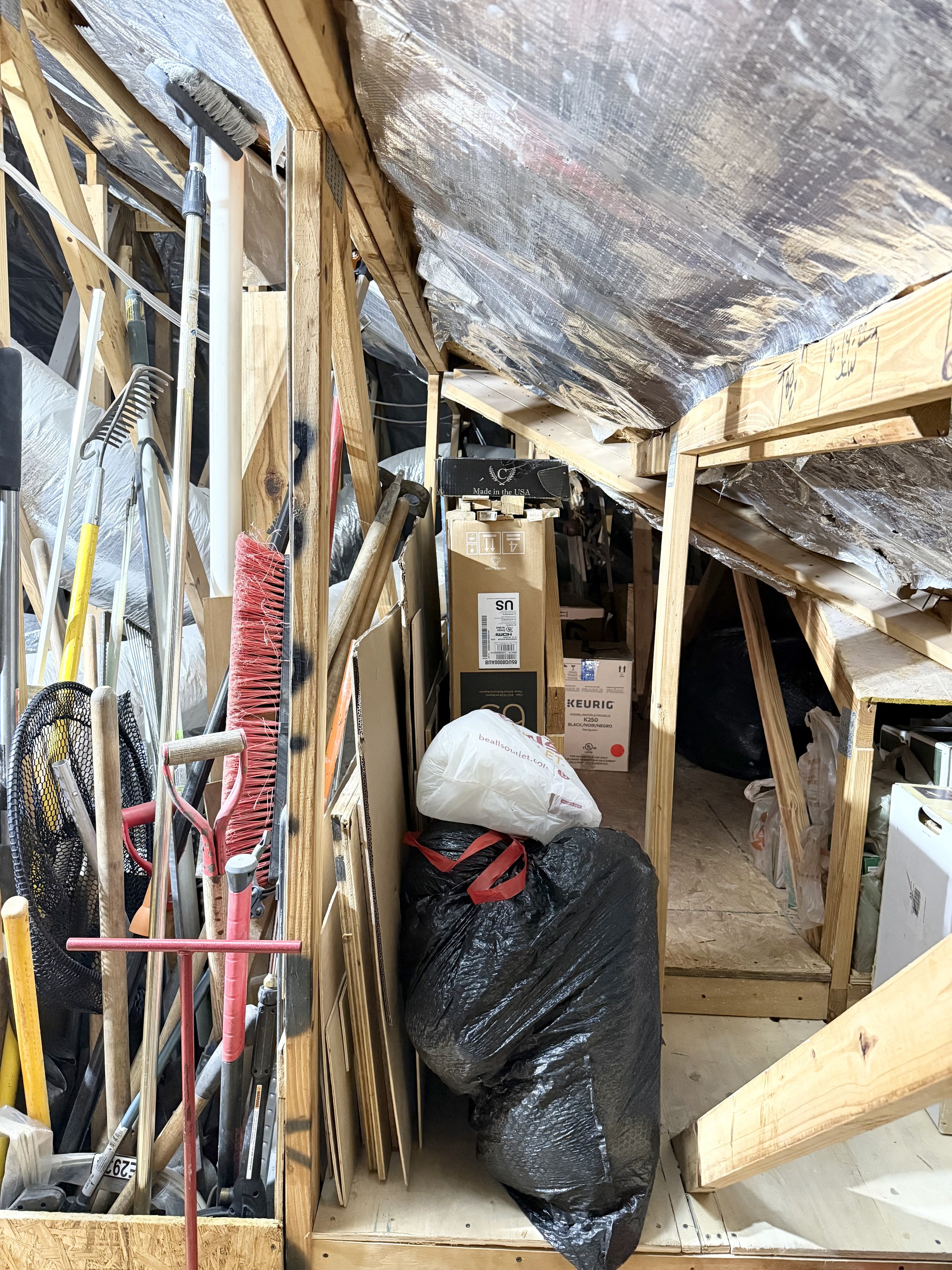 Attic storage space with various tools and boxes. Brooms and rakes are visible, along with cardboard boxes and a black garbage bag. Wooden framework supports the ceiling.