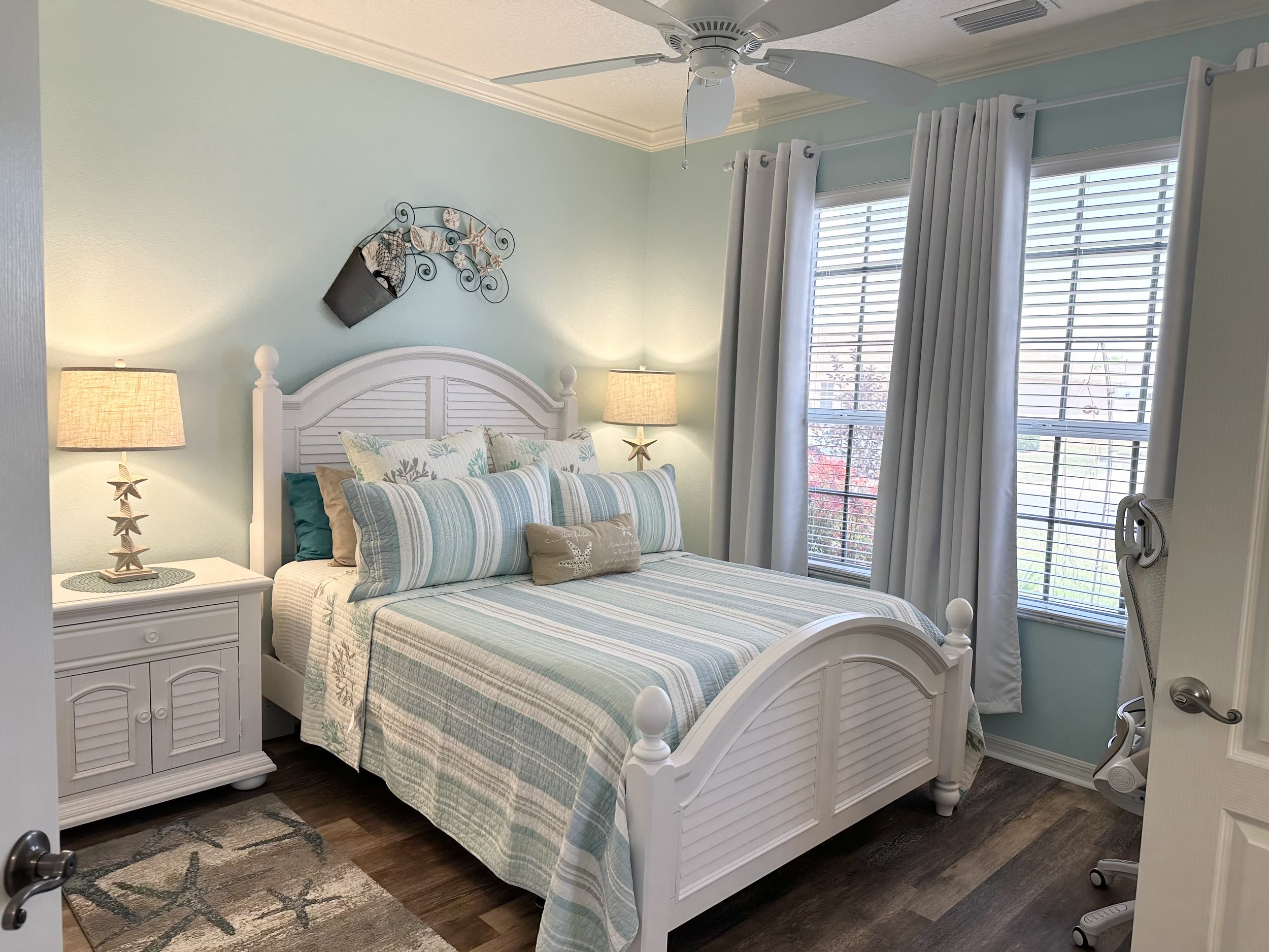Coastal-themed bedroom with white furniture and blue accents. Features include a bed with striped bedding, two bedside lamps with starfish stands, decorative wall art, and a light blue wall. The room has hardwood floors and two windows with white cur