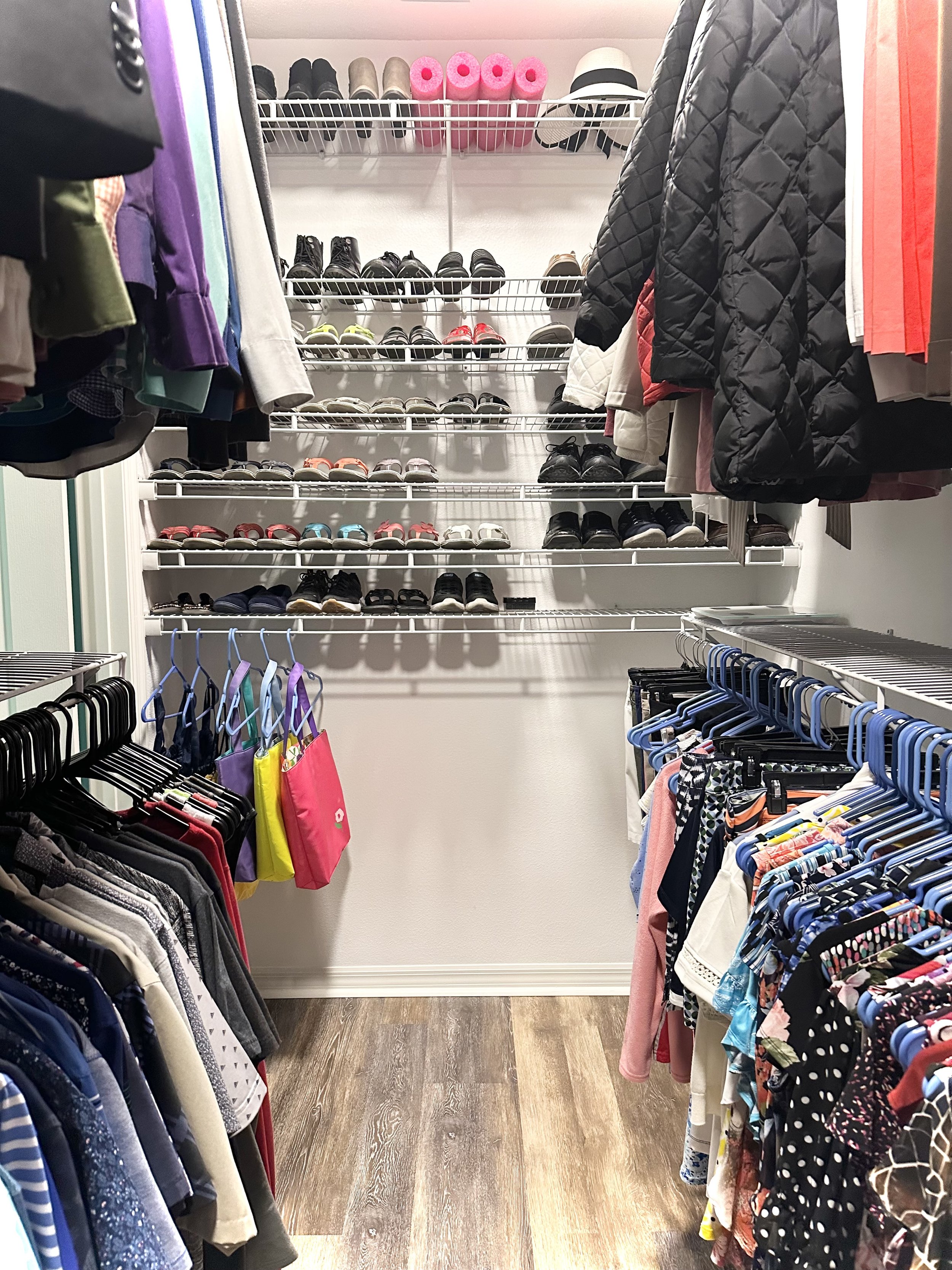 Organized walk-in closet with clothes, shoes, and accessories neatly arranged. Wire shelving holds shoes and hats, while colorful shirts and jackets hang on both sides. Tote bags are hung on hooks along the left side.