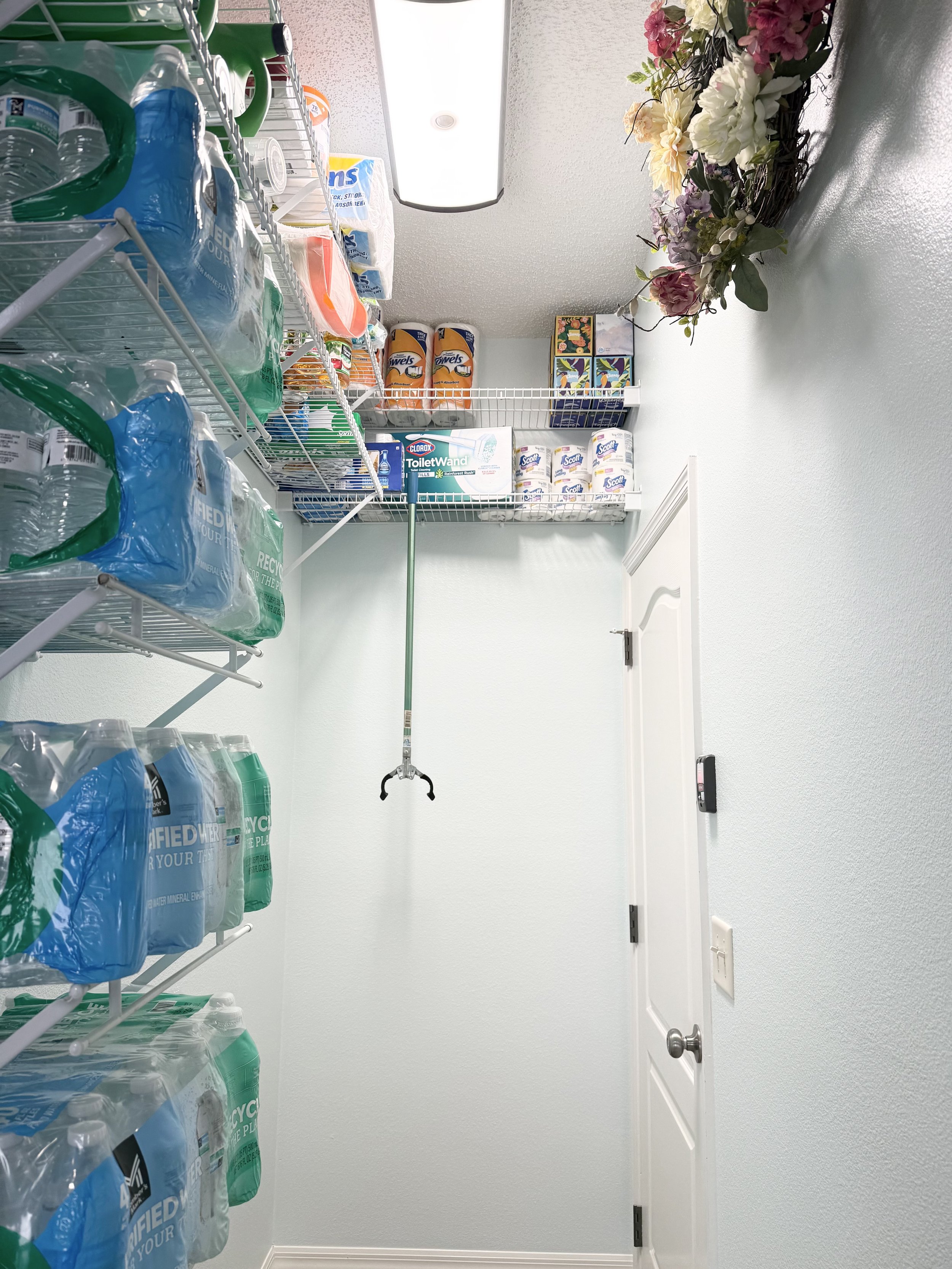 Storage room with shelves holding bottled water, paper towels, cleaning products, and decorative flowers hanging on the wall.