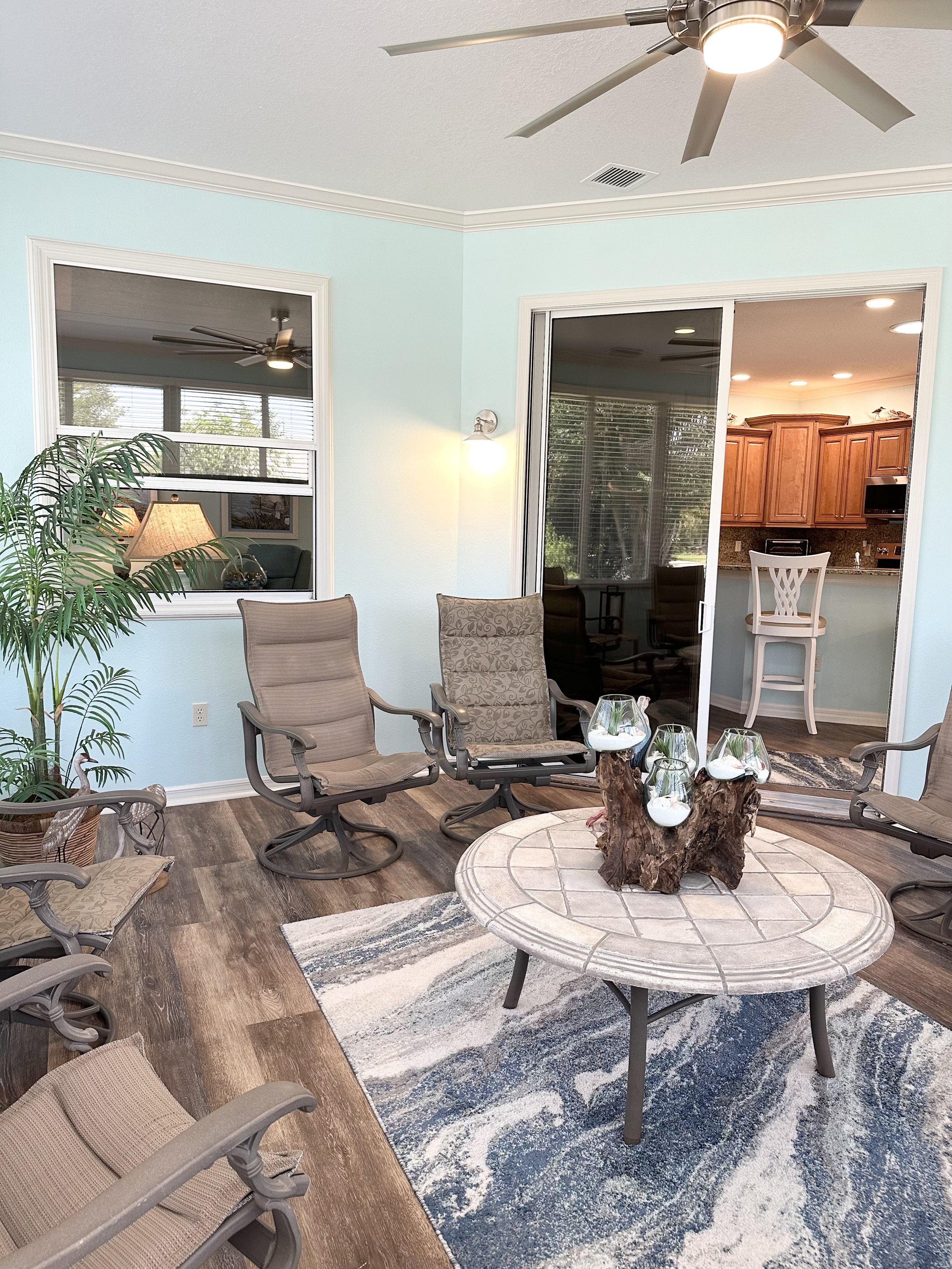 A sunroom with light blue walls featuring a ceiling fan and recessed lighting. It contains several cushioned patio chairs around a circular tile-top table with decorative glass vases on a wooden centerpiece. The flooring is wood-style and there's a b