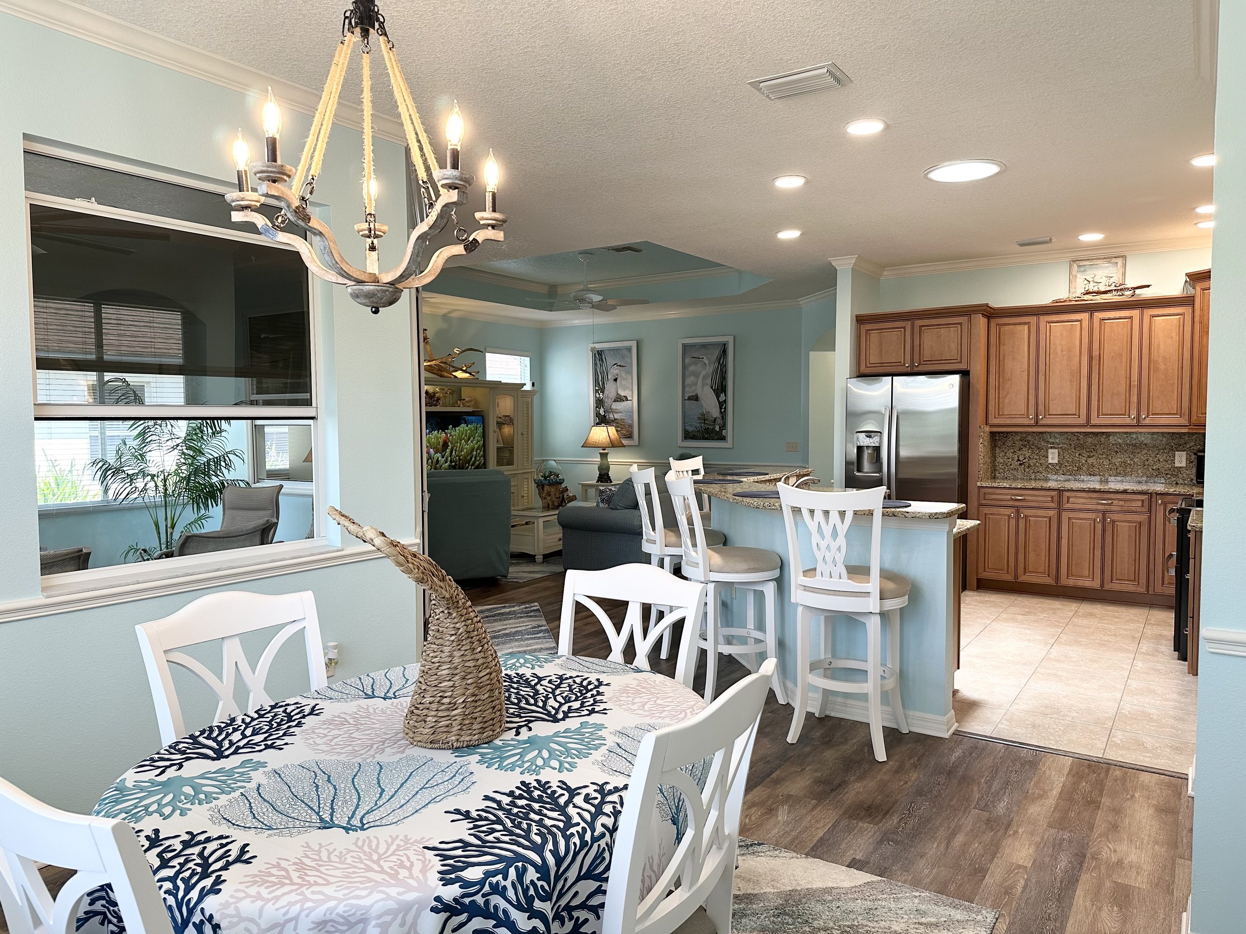 Beach-themed open-concept living space with dining area featuring white chairs and ocean-themed tablecloth, kitchen with wooden cabinets, granite countertops, bar stools, and a decorative chandelier.