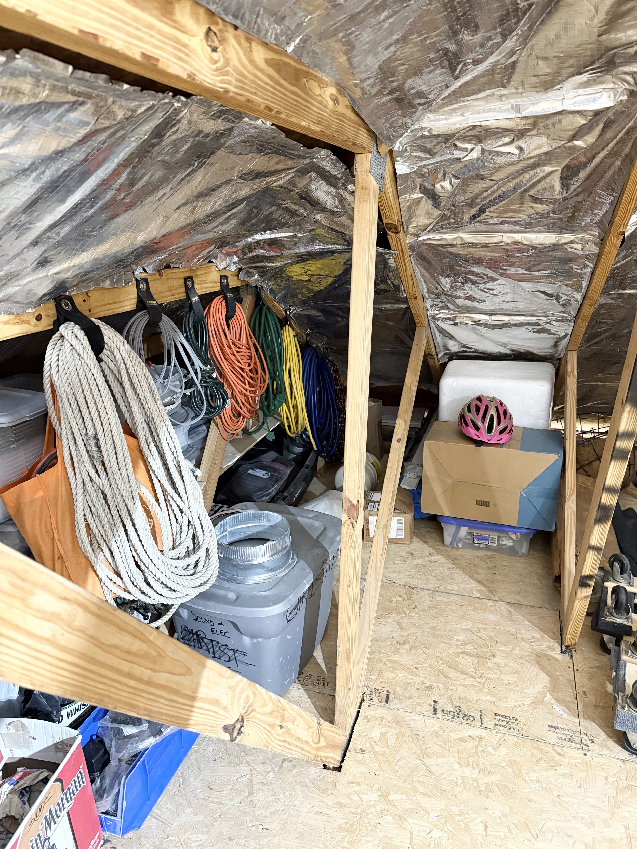 Attic storage space with exposed wooden beams and metallic insulation. Various colored extension cords are hanging on hooks, with ropes, bins, cardboard boxes, and a pink bicycle helmet visible. The floor is made of plywood.