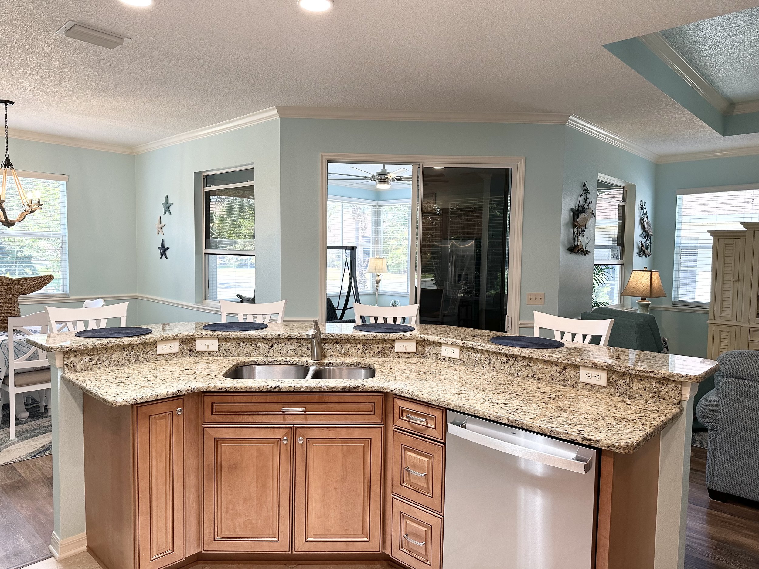 Modern kitchen interior with granite countertops, wooden cabinetry, and stainless steel sink and dishwasher. The room features bar seating with white chairs and blue place mats. Adjacent dining area visible with chandelier and decorative starfish wal