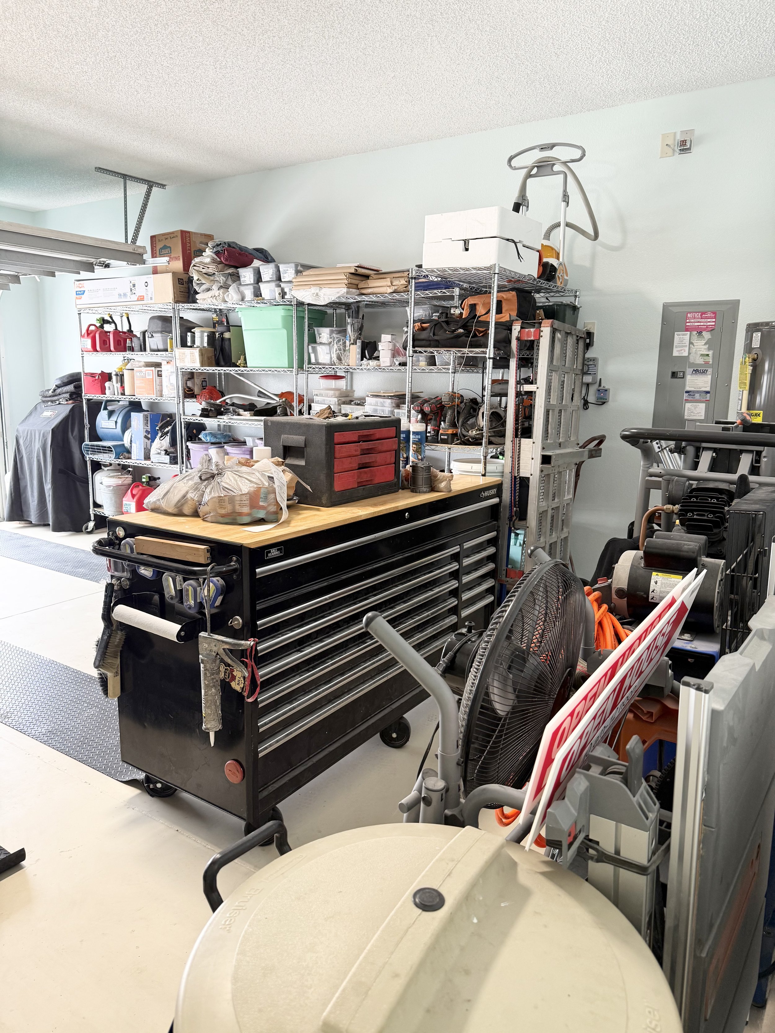 A cluttered garage with shelves filled with various tools, boxes, and containers. A large black tool chest with multiple drawers is in the center of the image. There's a fan, a large cooler, and an exercise machine visible. The space includes an asso