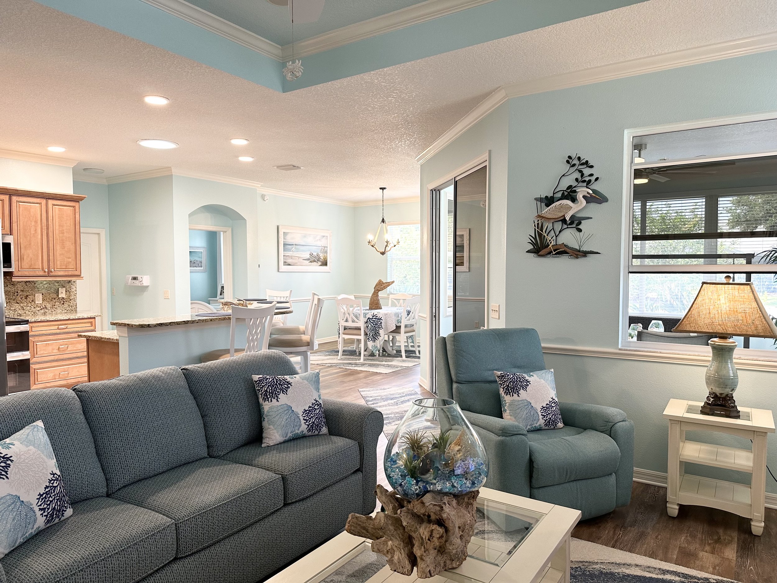 Coastal-style living room with blue sofas, decorative pillows, wooden accents, and a glass fishbowl centerpiece. The room features light blue walls, a lamp on a side table, wall art of a heron, and an adjoining kitchen area with a breakfast bar and d