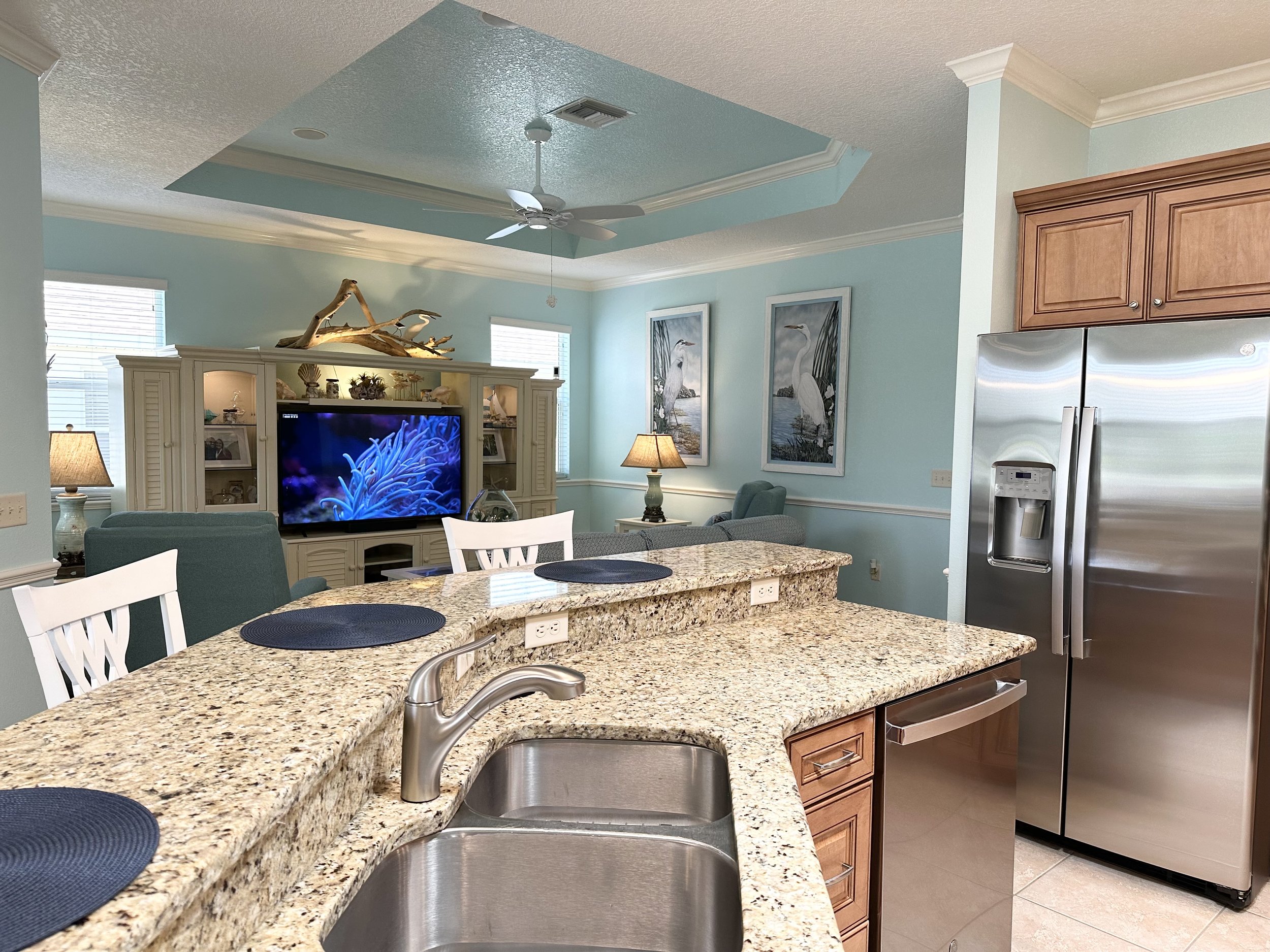 Modern kitchen and living room with granite countertops, double sink, stainless steel fridge, bar stools, wall-mounted TV, artwork, and decorative lighting.
