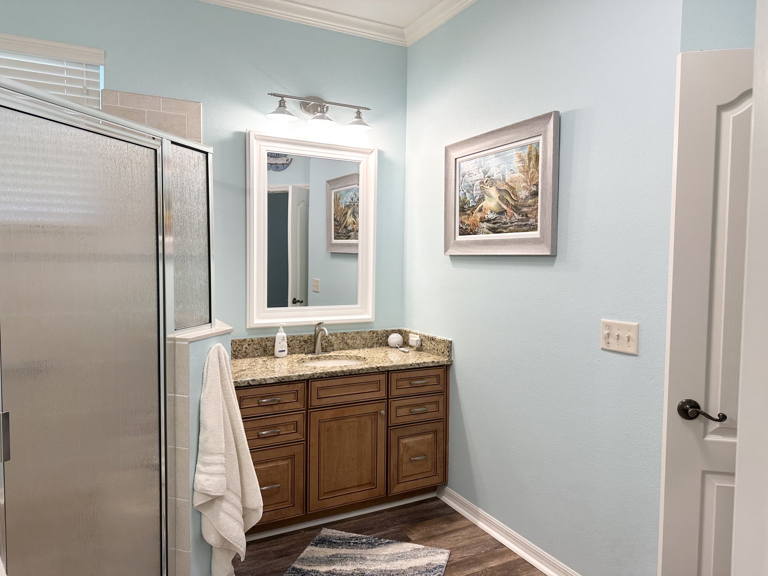 Bathroom with light blue walls, wooden vanity with granite countertop, wall mirror, framed artwork, glass shower enclosure, and towel hanging.