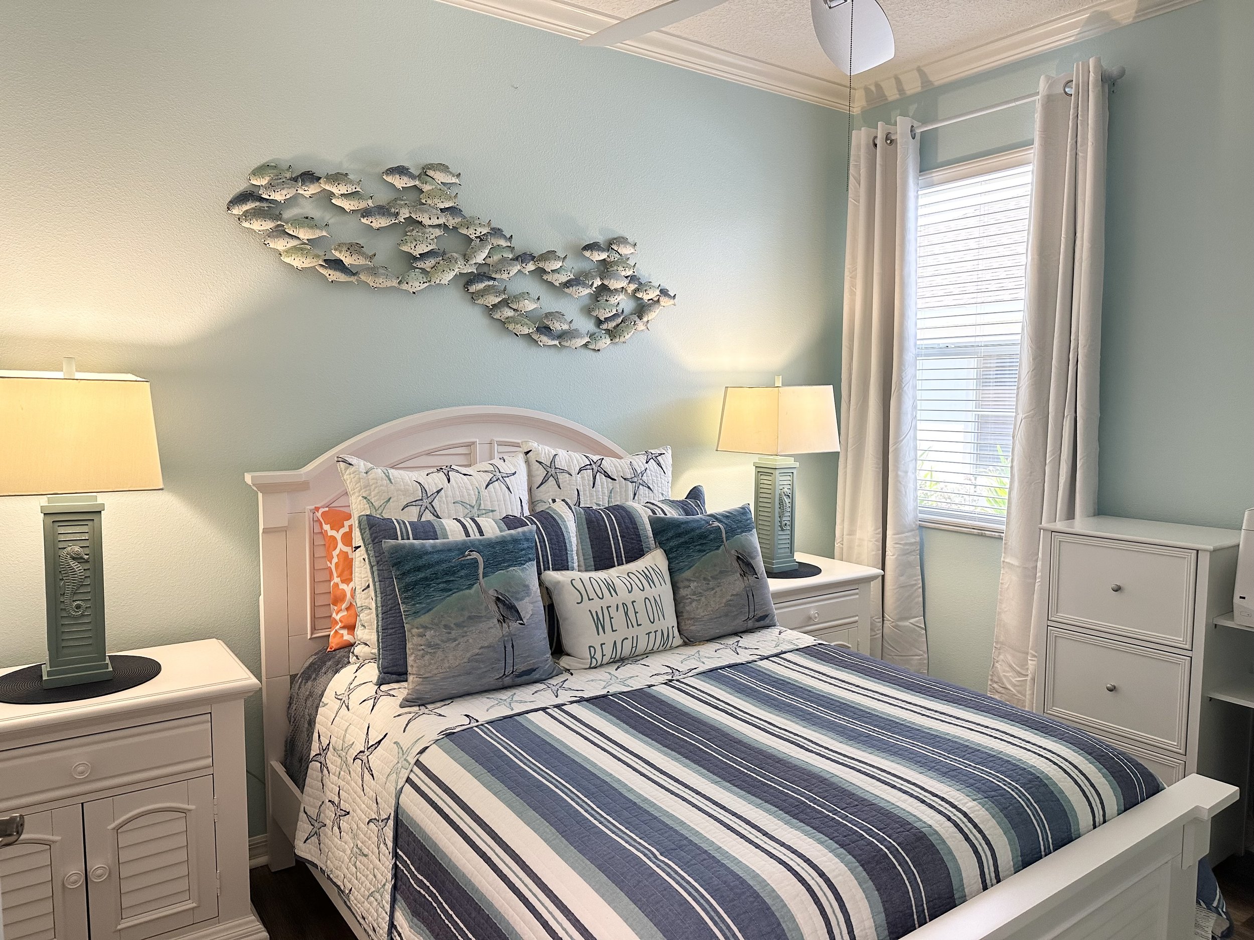 Coastal-themed bedroom with a white bed, blue striped bedding, decorative pillows, two side lamps, wall art of fish, and a window with curtains.