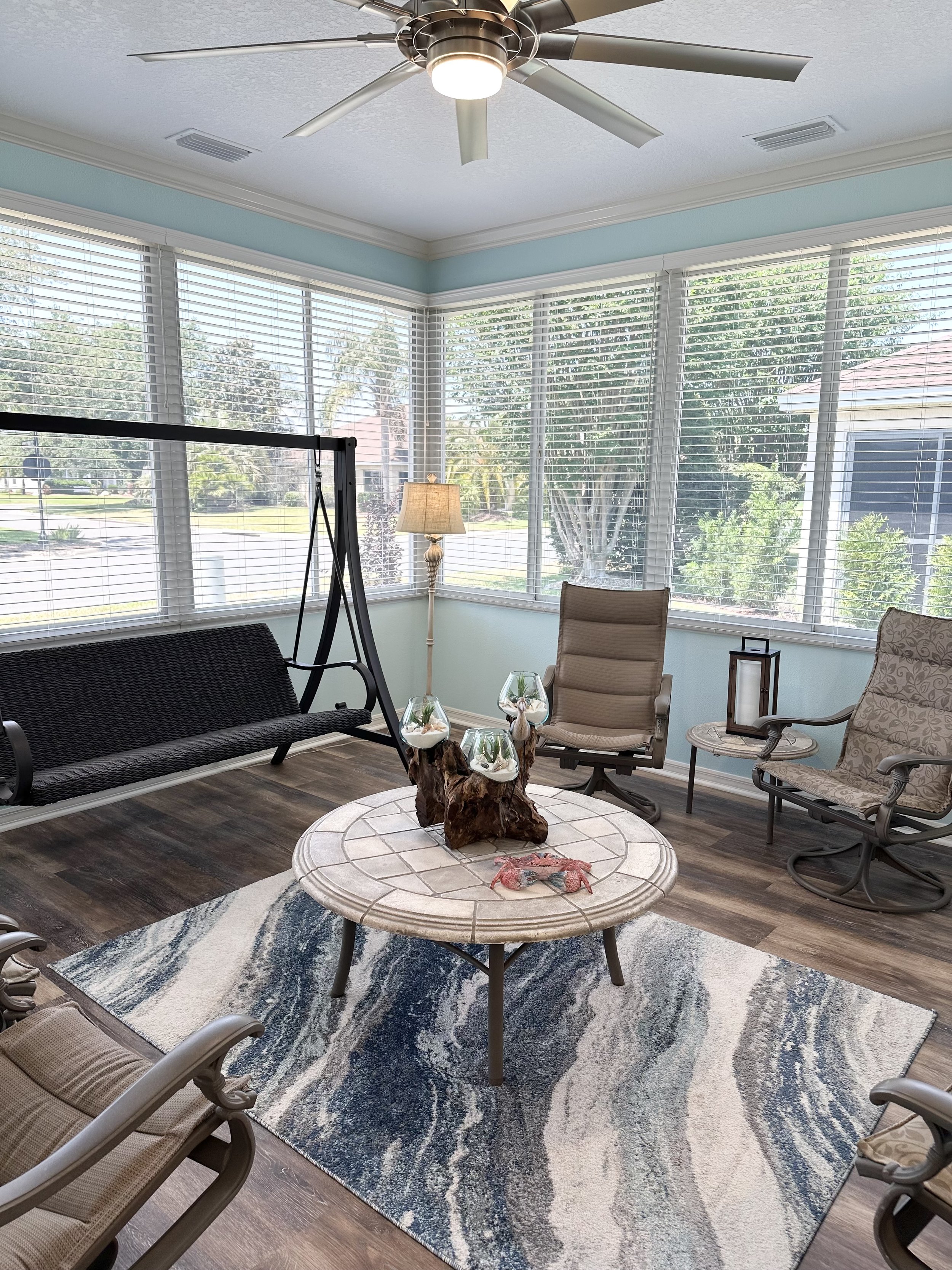Bright sunroom with ceiling fan, large windows, wicker swing, modern chairs, round table with decorative centerpiece, patterned rug, and floor lamp.