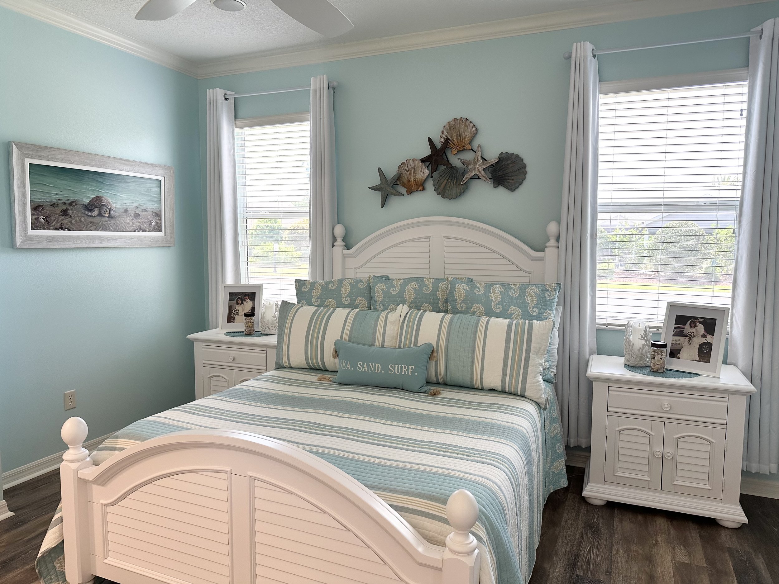 Beach-themed bedroom with a white bedframe, striped blue and white bedding, and decorative pillows. There are two nightstands with photos and decor items, and a seashell wall art piece above the bed. Light blue walls, a ceiling fan, and sheer white c