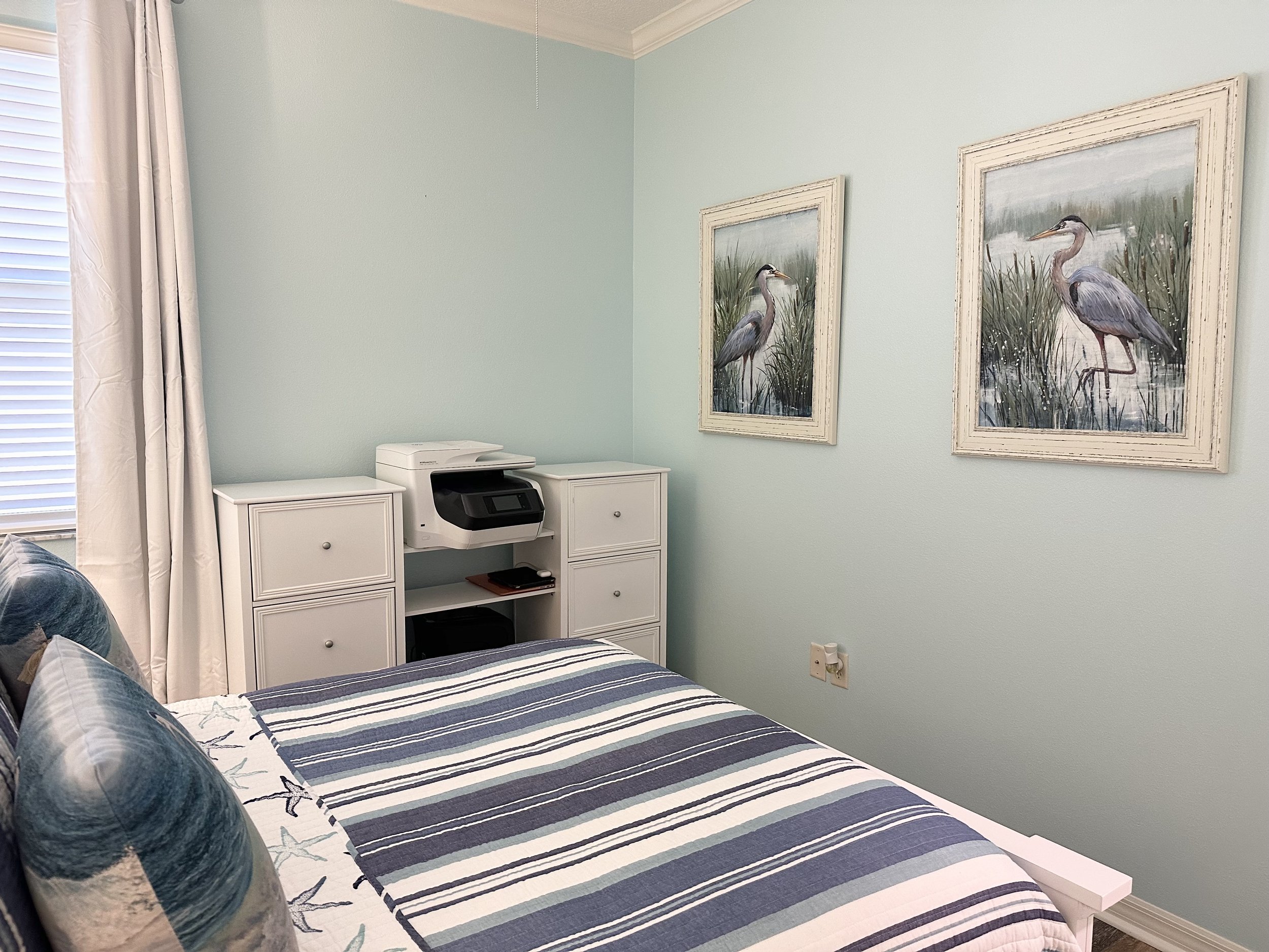 Bedroom with light blue walls, striped bedding, two framed bird paintings, and a printer on a white storage unit.
