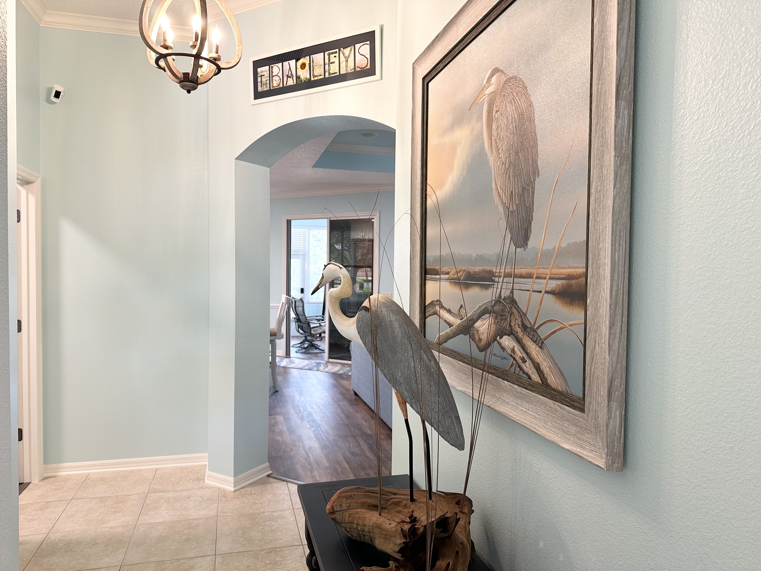 Interior hallway with light blue walls, a chandelier, and a heron sculpture next to a framed heron artwork. A sign reading 'The Baileys' is above an archway leading to another room with a visible desk and chair.