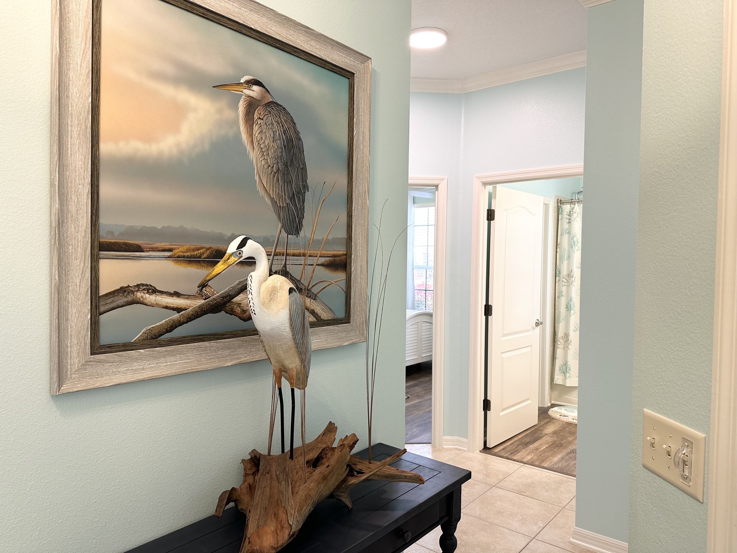 Interior of a hallway with light blue walls, featuring a framed painting of a heron and a heron sculpture on a table. The hallway leads to a room with an open door, revealing a glimpse of a bathroom with a shower curtain.