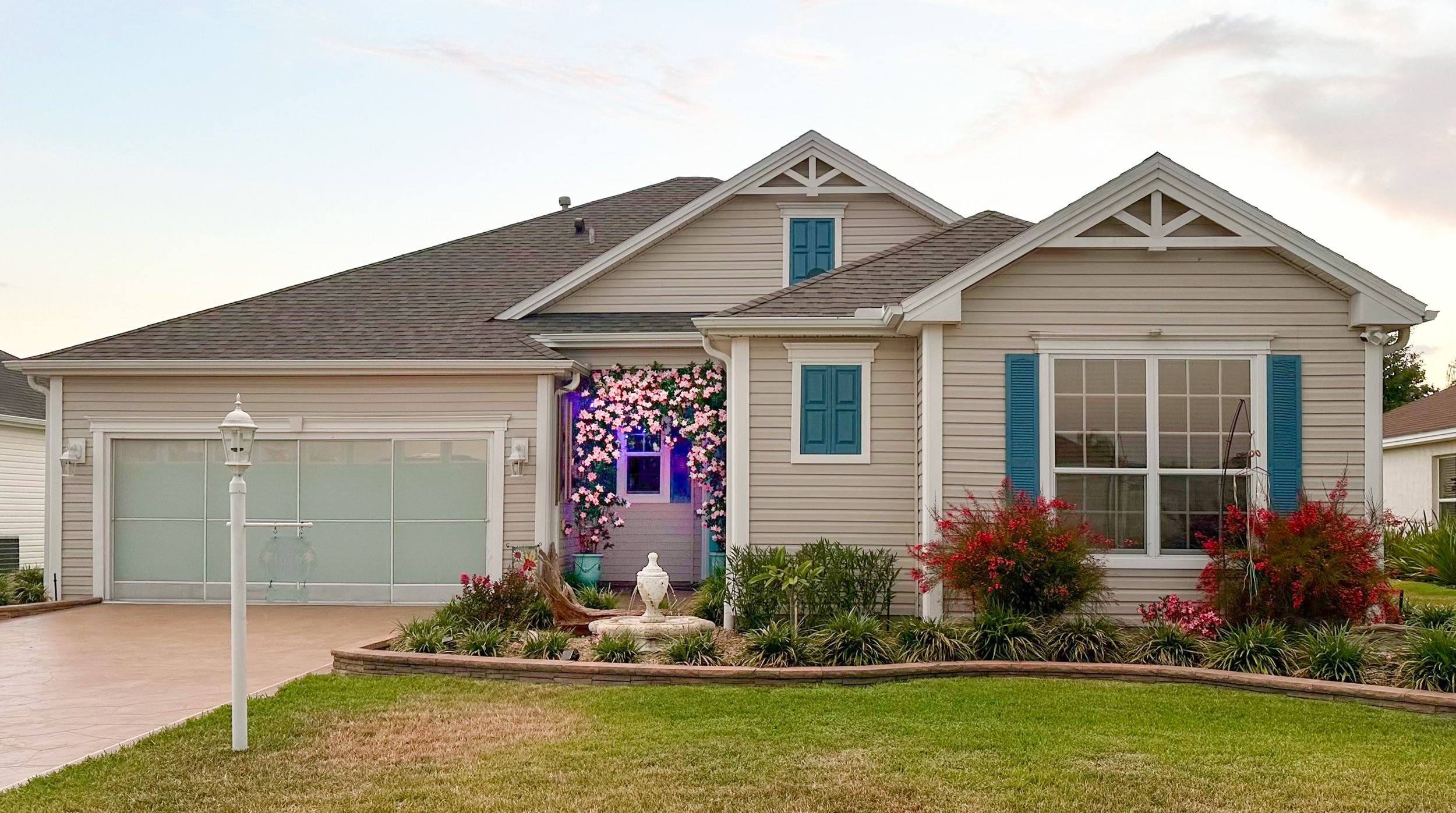 Front view of 1676 Van Buren Way, The Villages, FL. This house has a well-maintained lawn and garden, featuring pink flowers, a white birdbath, and a small lamppost. The garage has security screen covering the main garage door.