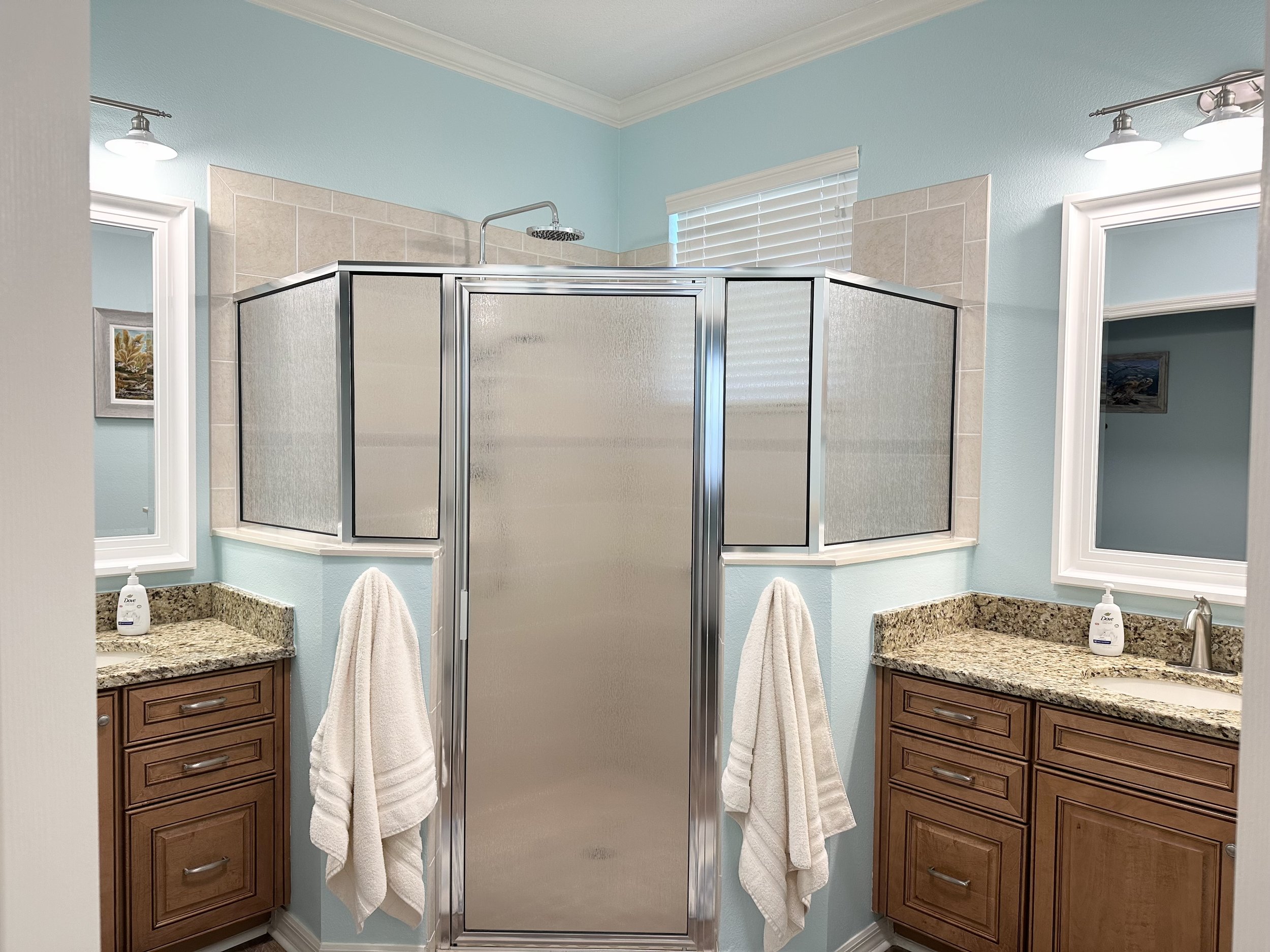Modern bathroom with a glass shower, dual sinks with granite countertops, wooden cabinets, and wall-mounted mirrors. Light blue walls and artificial lighting complete the decor.