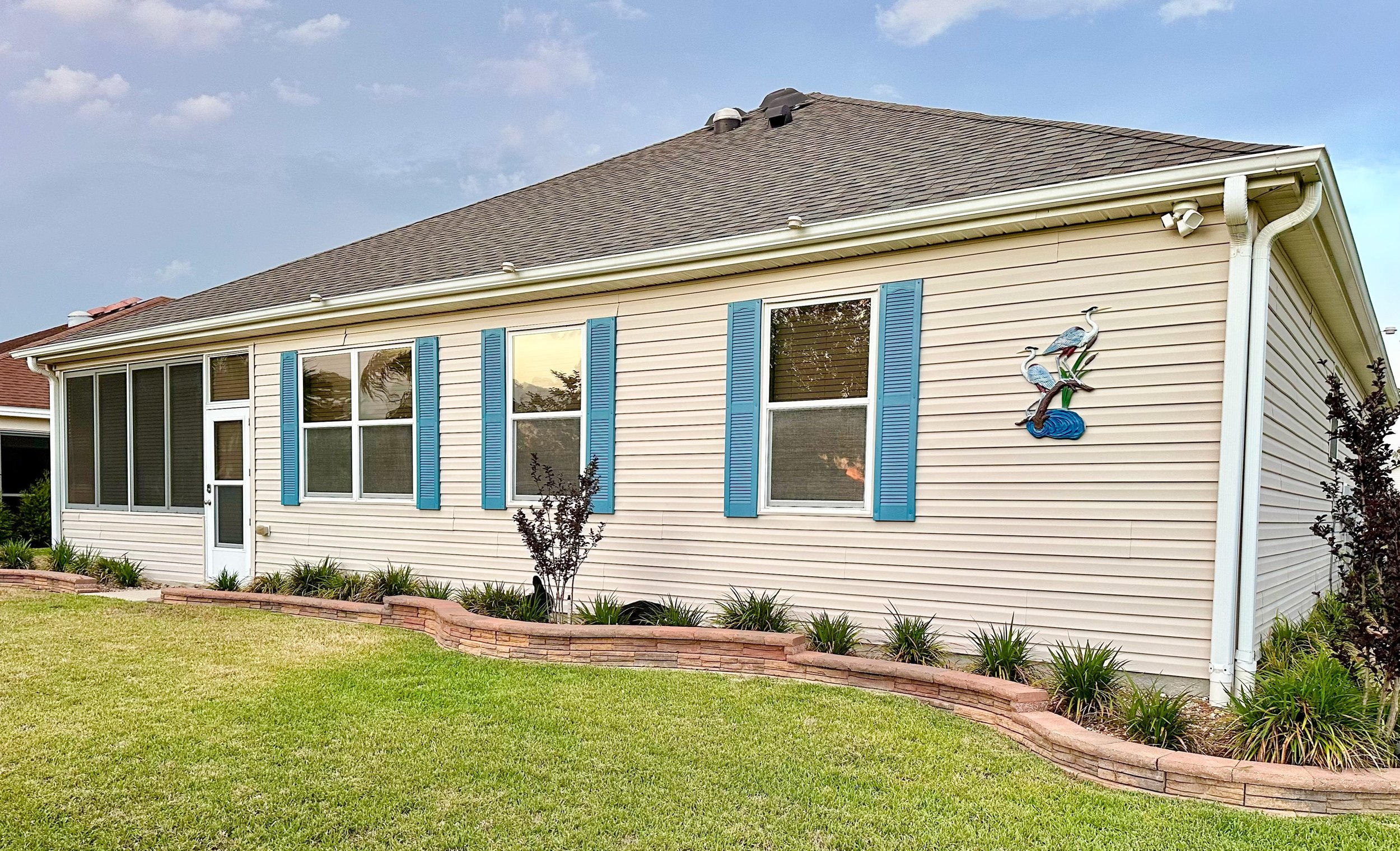 A single-story house with beige siding and blue shutters, featuring three large windows and a door on one side. The house has a neatly manicured lawn and a brick-edged flower bed with small plants. Decorative bird art is mounted on the exterior wall.