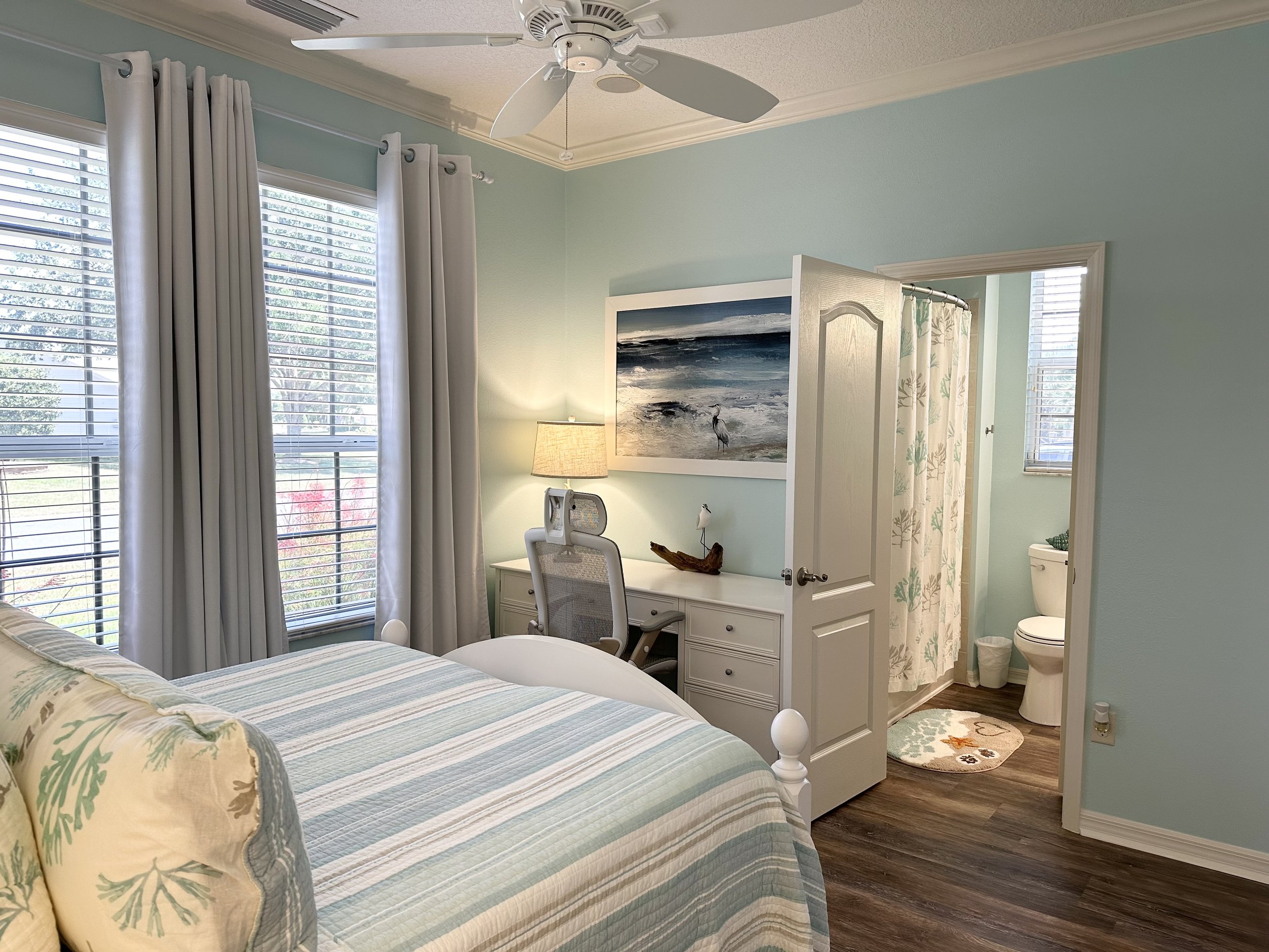 Coastal-themed bedroom with a light blue and white color scheme. Features a bed with striped bedding, a desk with a chair, a lamp, and an ocean-themed picture on the wall. An open door leads to a small bathroom with a floral shower curtain and a wind