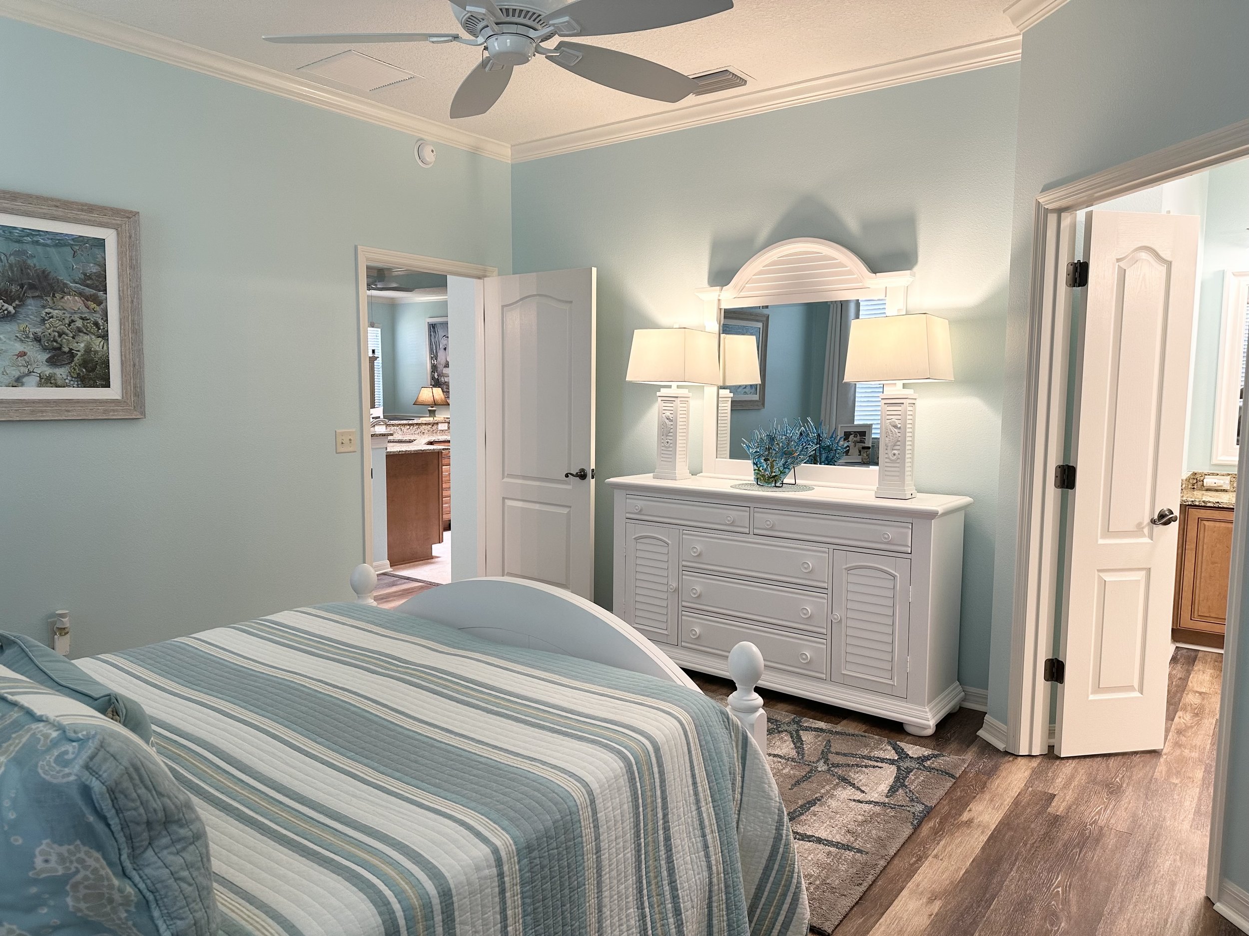 A bedroom with light blue walls and wooden flooring features a bed with a striped comforter. A white dresser with a mirror and two lamps stands against one wall. The room has a ceiling fan and has a partially open door leading to a bathroom, visible