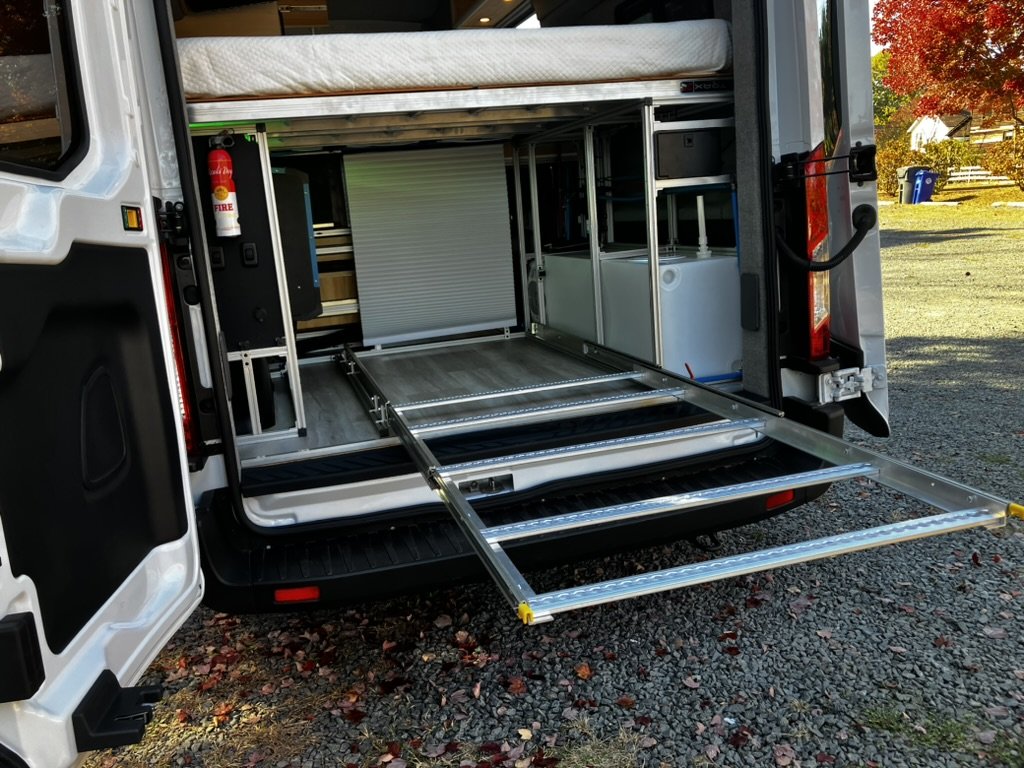 Open vehicle with a fold-out metal ramp, interior shelving, and a fire extinguisher, parked outdoors on a gravel surface with trees and a blue trash bin in the background.