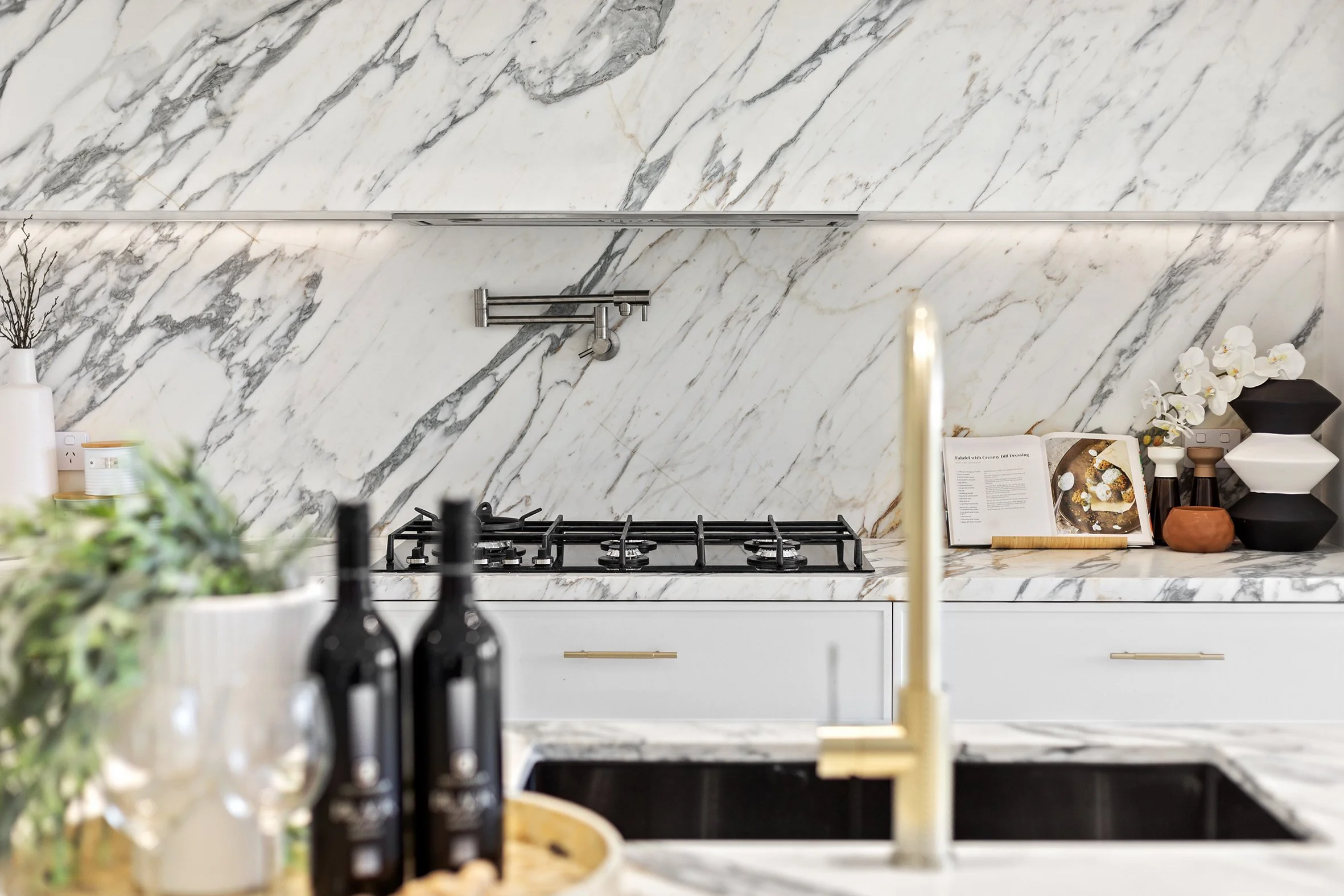 Modern kitchen with white marble backsplash, countertop, and black gas stovetop. Decor includes black bottles, a tray, candles, and an open cookbook, with white and black vases and orchids on the counter.